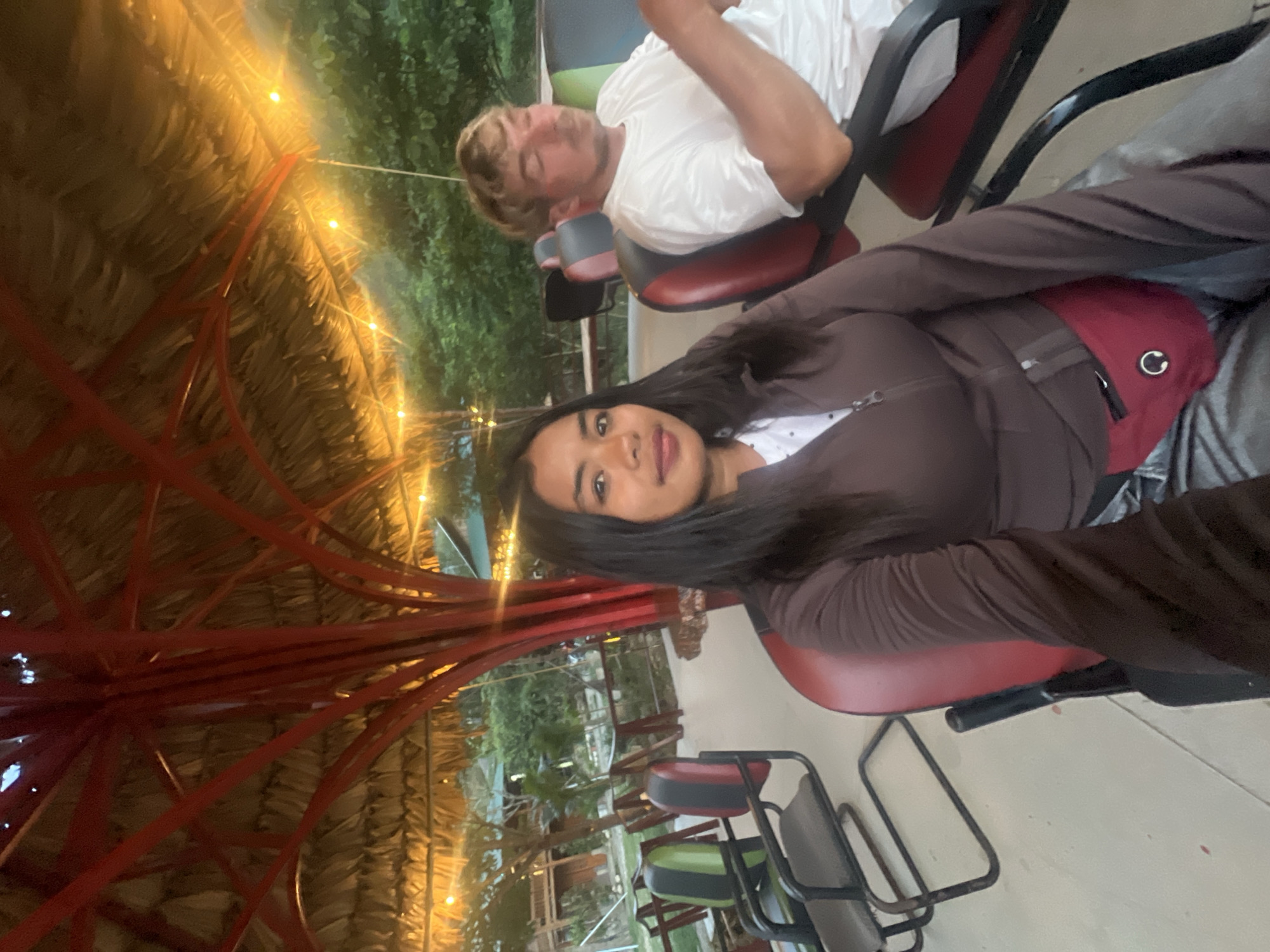 In the city of Tu Le, Vietnam, a young woman with long, dark hair and a brown zip-up jacket is seated in the foreground. She wears a red fanny pack across her waist and a white shirt underneath. To her right, a man with blond hair and a white t-shirt is also seated, looking downwards. They are positioned under a thatched roof supported by a red, branching structure. String lights are visible above them. In the background, lush green foliage and trees can be seen, with glimpses of wooden structures and pathways suggesting an outdoor seating area or resort. The lighting indicates it is either dusk or early evening, with artificial lights adding warmth to the scene. The seating arrangement consists of chairs with black frames and red, black, and green upholstery. The ground is paved and appears to be a light concrete color.