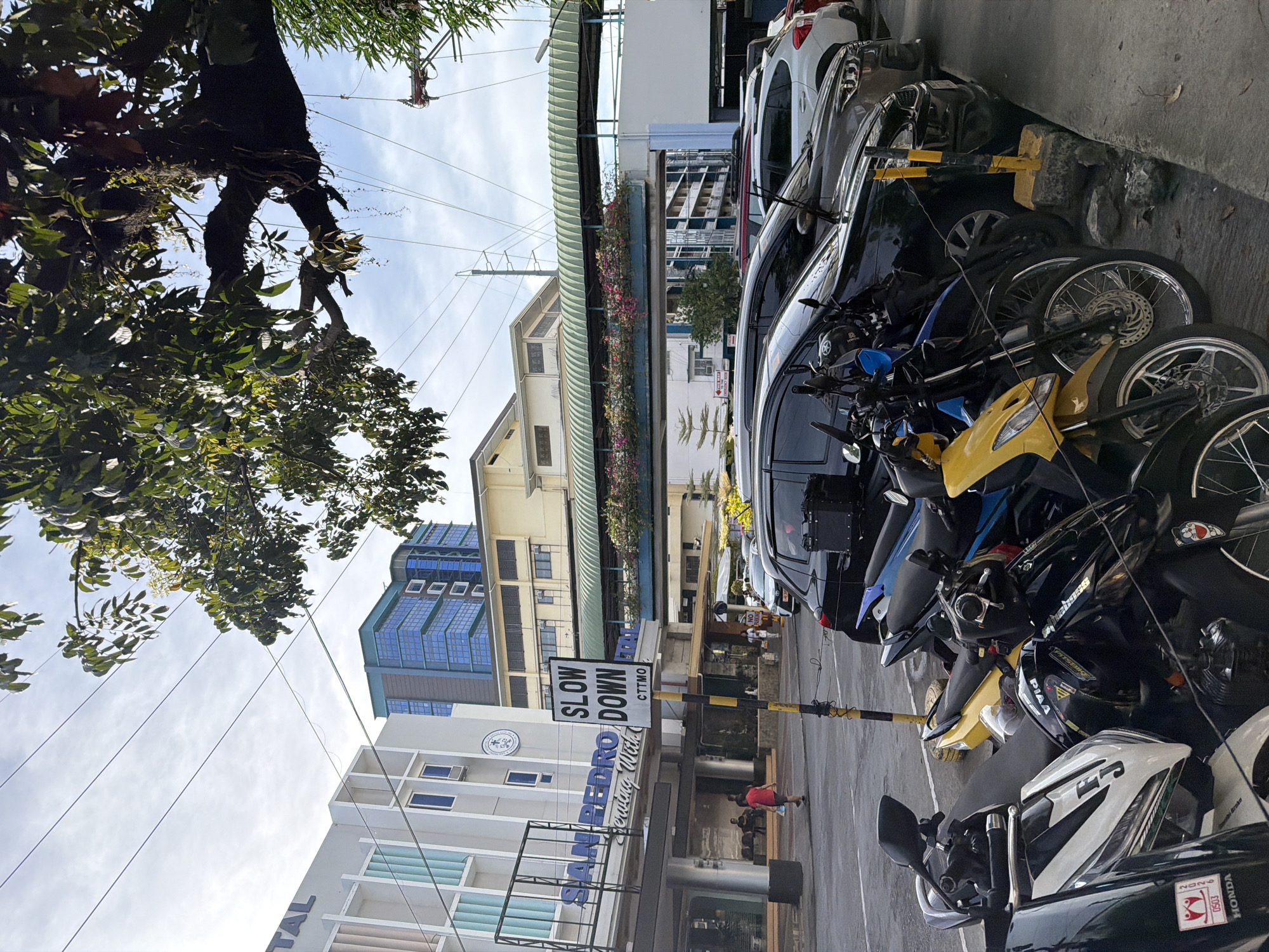 The image depicts a street scene in Davao City, Philippines. A building with the name "SAN PEDRO Hospital" is visible. A blue building is also present. A "SLOW DOWN CTTMO" street sign is present. Several motorcycles and cars are parked along the side of the road. A tree with dense foliage is visible in the upper left corner of the image. Two people, including a person wearing red, are present in the cross walk.