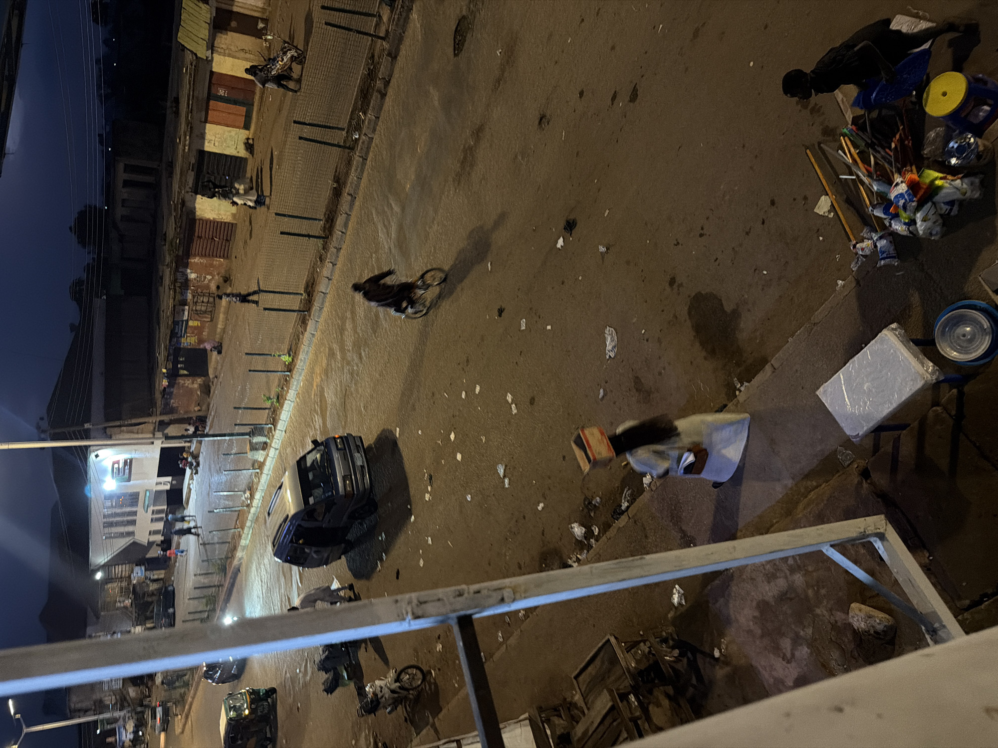 The image depicts a street scene in Bukuru, Nigeria, at dusk. In the foreground, a woman walks on the sidewalk, carrying a box on her head and wearing a white robe. A white metal railing is visible on the left. The street is paved and appears somewhat worn. Several vehicles are present, including a blue van, a tricycle, and a motorcycle. People are walking and riding bicycles on the street and sidewalk. Shops with closed doors line the street, some with colorful advertisements. Debris and scattered paper are visible on the ground. In the mid-ground, a person is riding a bicycle, casting a long shadow. Further down the street, two individuals are seen pushing a cart filled with items. The overall atmosphere is one of a bustling but somewhat untidy urban environment.