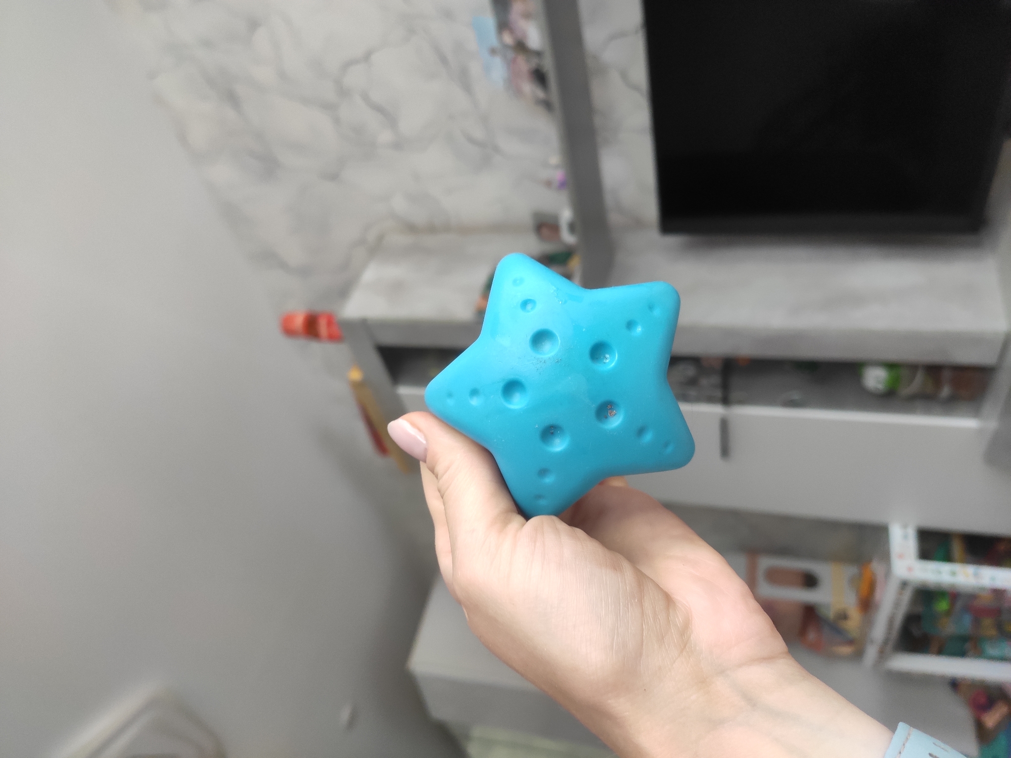 A person is holding a blue, star-shaped object with small, circular indentations on one side in their hand. The person's hand is fair-skinned with manicured nails. The background includes a blurred TV and a light gray and white marble-patterned wall behind a gray storage unit. The environment suggests the interior of a house or apartment. The city is not explicitly mentioned but assumed to be Sterlitamak, Russia.