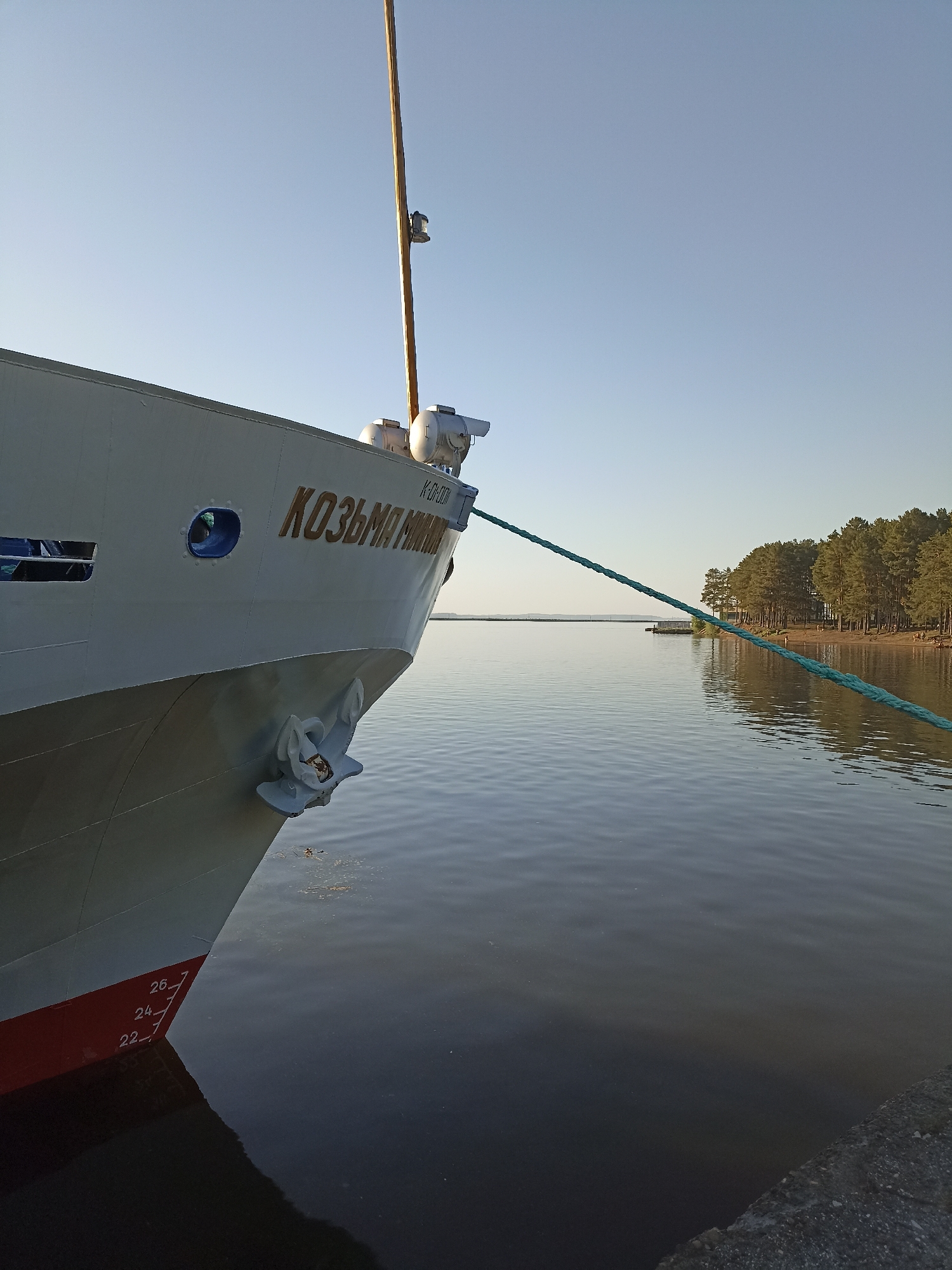The image shows the bow of a ship named "KOZ'MA MININ" docked at an unspecified location in the city of Unknown, Unknown. The ship is grey with red markings near the waterline. A green rope stretches from the bow towards a distant shoreline lined with trees. The water is calm. The sky is clear and light blue. An anchor is visible on the bow. A pole extends upwards from the bow.