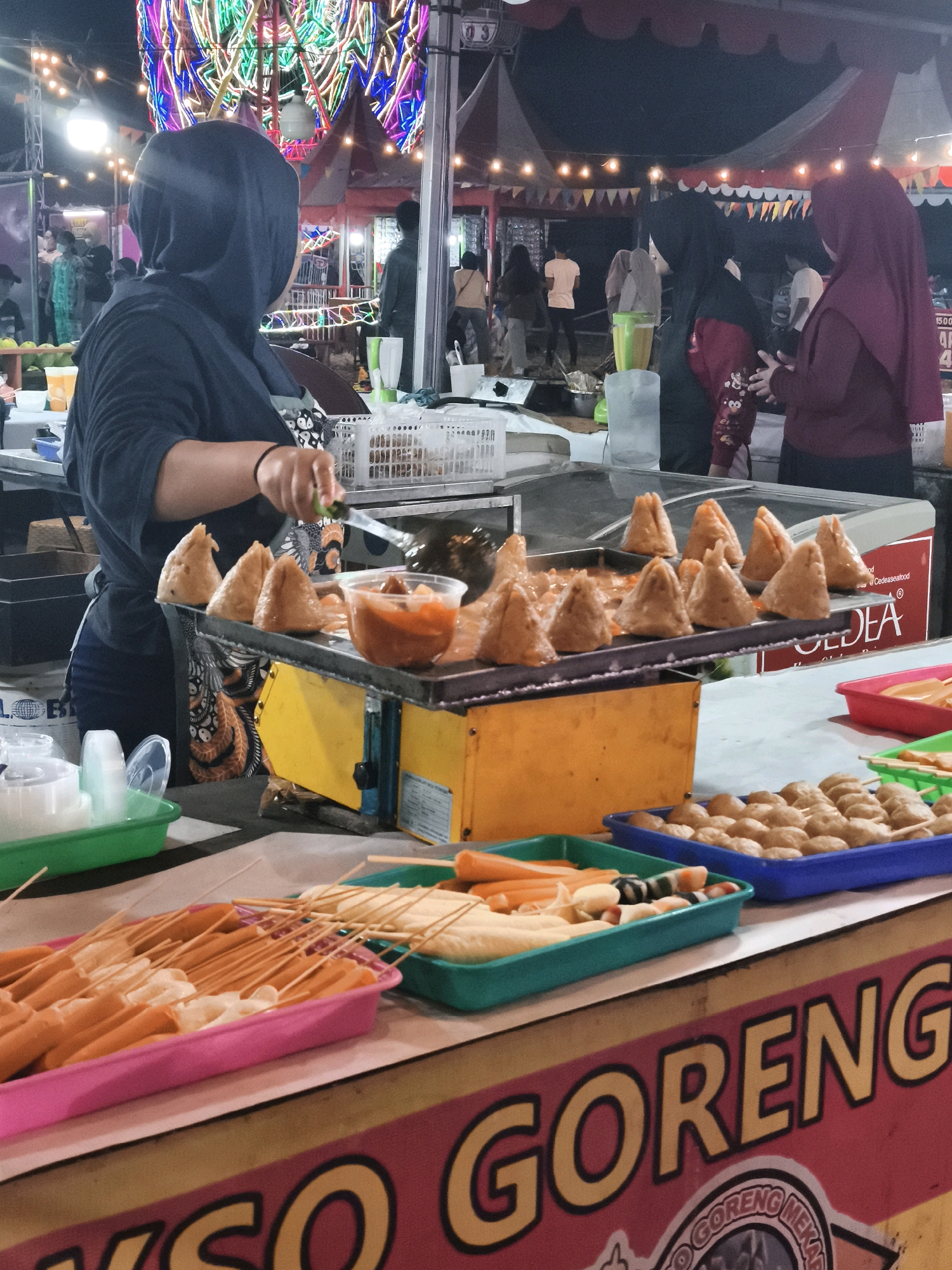 The image depicts a nighttime scene at an outdoor food stall, possibly in Palembang, Indonesia, given the "GORENG" sign which translates to "fried" and is a common word on Indonesian food stalls. In the foreground, a woman wearing a dark hijab and an apron is actively preparing food. She is holding a knife over a tray of triangular-shaped fried items, some of which appear to be stuffed. Adjacent to this tray is a bowl of dipping sauce. In front of the cooking area, several plastic trays hold various skewered food items, including sausages and what appear to be meatballs. These are arranged on the counter in front of the stall.
Behind the vendor, other people are visible, some wearing headscarves. In the background, a brightly lit Ferris wheel, adorned with colorful lights, is a prominent feature, suggesting a festive or carnival-like atmosphere. String lights and banners further enhance the ambiance of the market or fair. The overall environment is that of a bustling night market or festival.