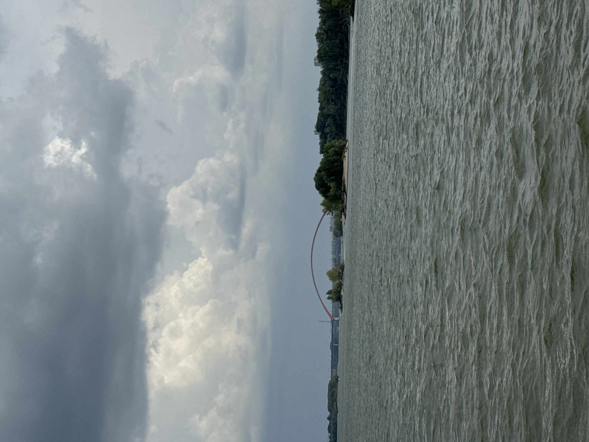 The image shows a wide body of water, likely the Ob River in Novosibirsk, Russia, under a cloudy sky. A long, arched red bridge spans the river in the distance, with smaller islands covered in trees visible in the foreground and middle ground. The water is choppy, indicating a breezy or rough conditions. The overall scene is somewhat overcast and moody, with dramatic clouds filling the sky.