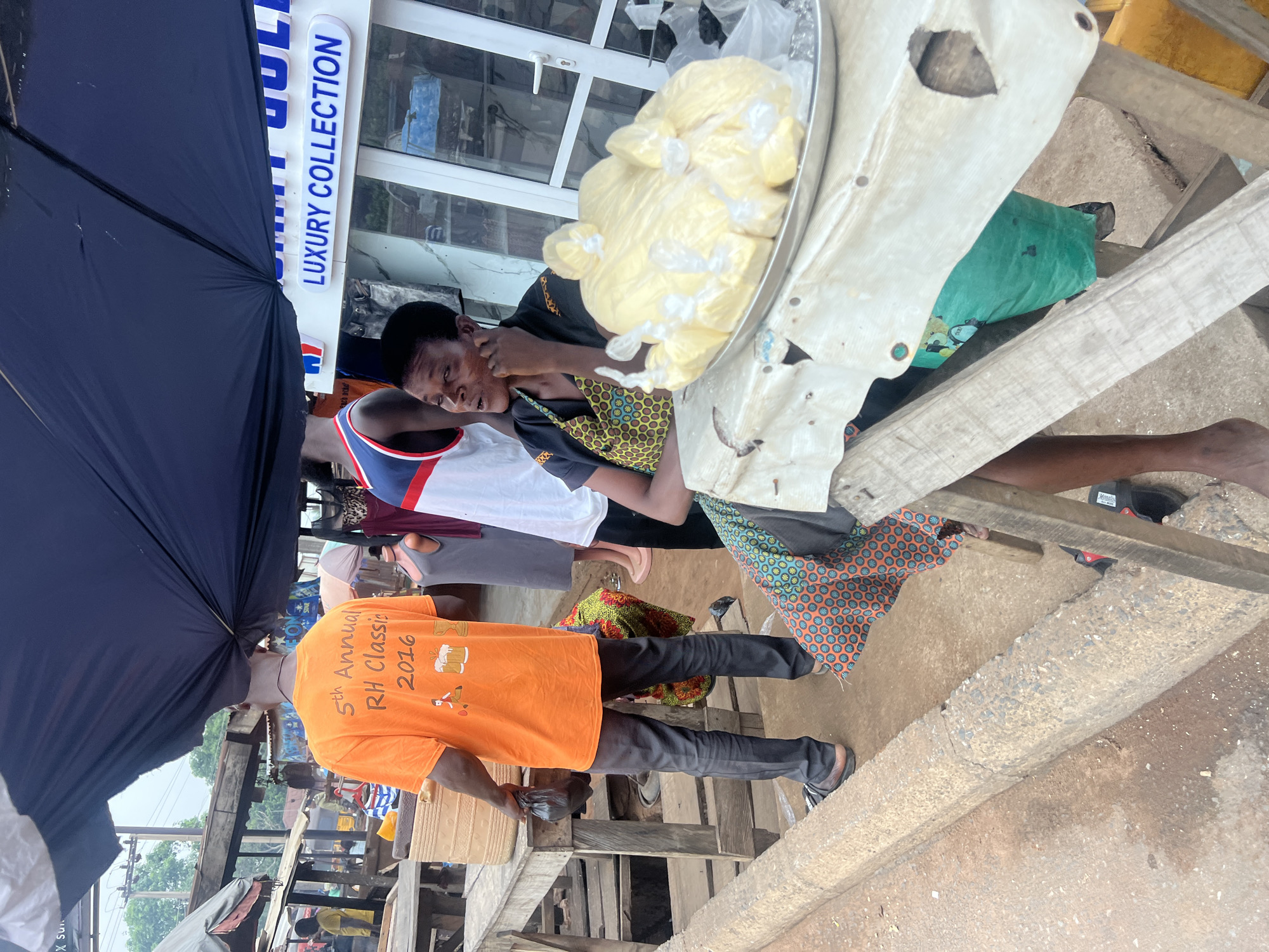 The image, taken in Obosi, Nigeria, shows an outdoor market scene. A woman wearing a black top and patterned apron sits behind a table, with a large, silver tray piled with bags of what appears to be shea butter. An individual in an orange t-shirt reading "5th Annual RH Classic 2016" stands facing away from the camera. Two other individuals are visible, with one in a white shirt and blue-and-red trim. A black umbrella covers part of the scene. The background shows a building with a sign reading "Luxury Collection." The ground appears to be a combination of concrete and dirt.