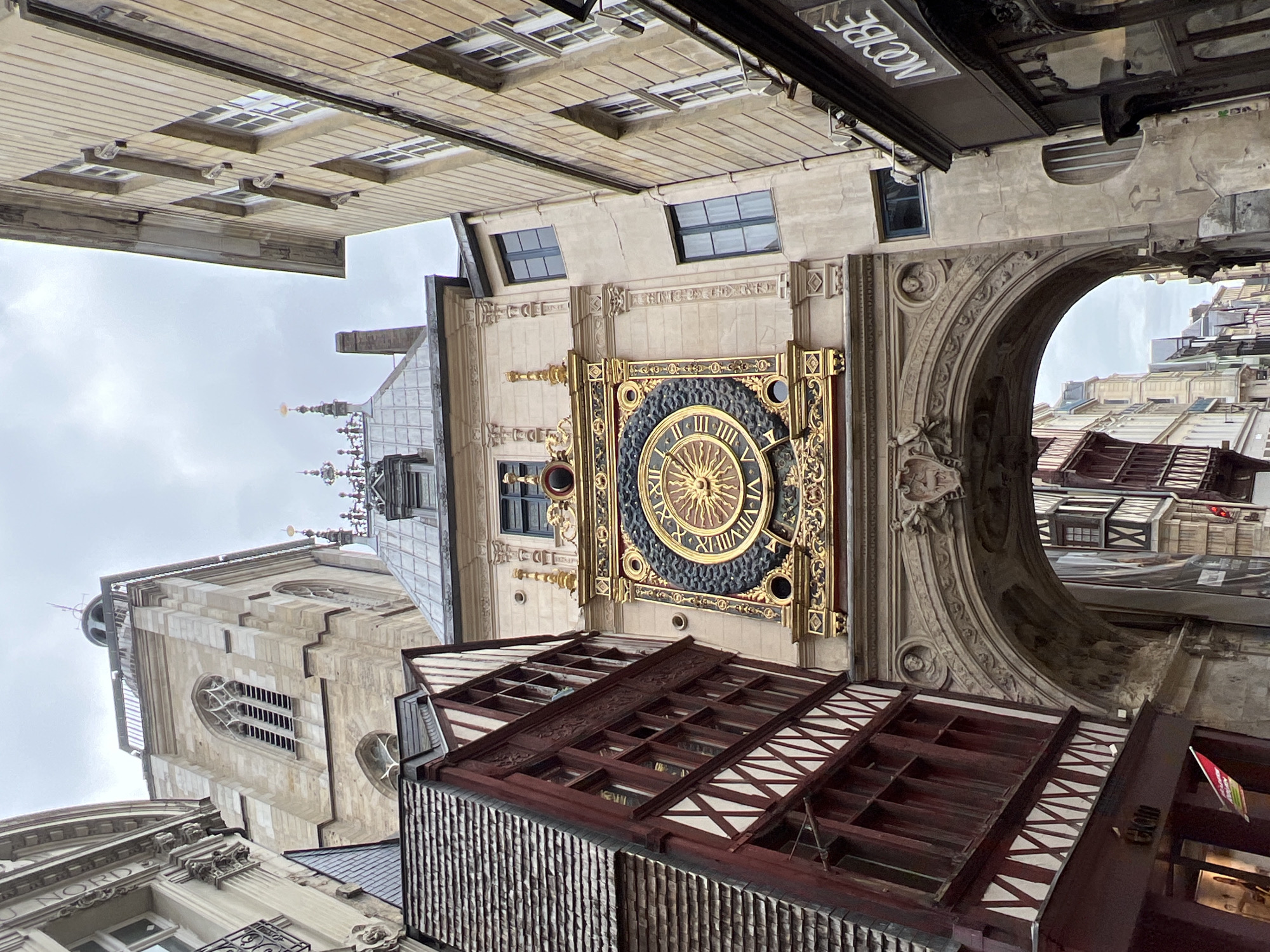 This media file captures a historic street scene in Rouen, France, dominated by the elaborate Gros Horloge (Great Clock). The image is taken from a low angle, looking up at the architectural details of the famous astronomical clock and its surroundings.
The central subject is the ornate golden clock face, featuring Roman numerals and a radiant sun motif at its center, set against a dark blue or black background. This clock is integrated into a larger stone building, beneath a grand archway that spans the street. The archway and the clock's frame are richly decorated with intricate carvings and sculptural elements.
To the left of the clock, a tall, light-colored stone belfry rises, characteristic of a church or civic tower, with gothic-style arched windows and decorative finials at its summit. Below the clock and extending into the foreground are traditional timber-framed buildings, displaying dark reddish-brown wood beams against white or light-colored infill panels, some with distinctive crisscross patterns. The roofs or upper sections of these buildings feature varied textures, including slatted wooden panels.
Through the archway on the right, more distant buildings, some also timber-framed, are visible, suggesting a narrow, winding street typical of an old European city. The sky above and through the archway is overcast, indicating a cloudy daytime setting.
Visible text includes a partially inverted sign above the archway that reads "GROS HORLOGE," and another inverted sign on a building to the lower left that says "DU NORD." No people or specific activities are depicted; the focus is entirely on the historical architecture and urban landscape.