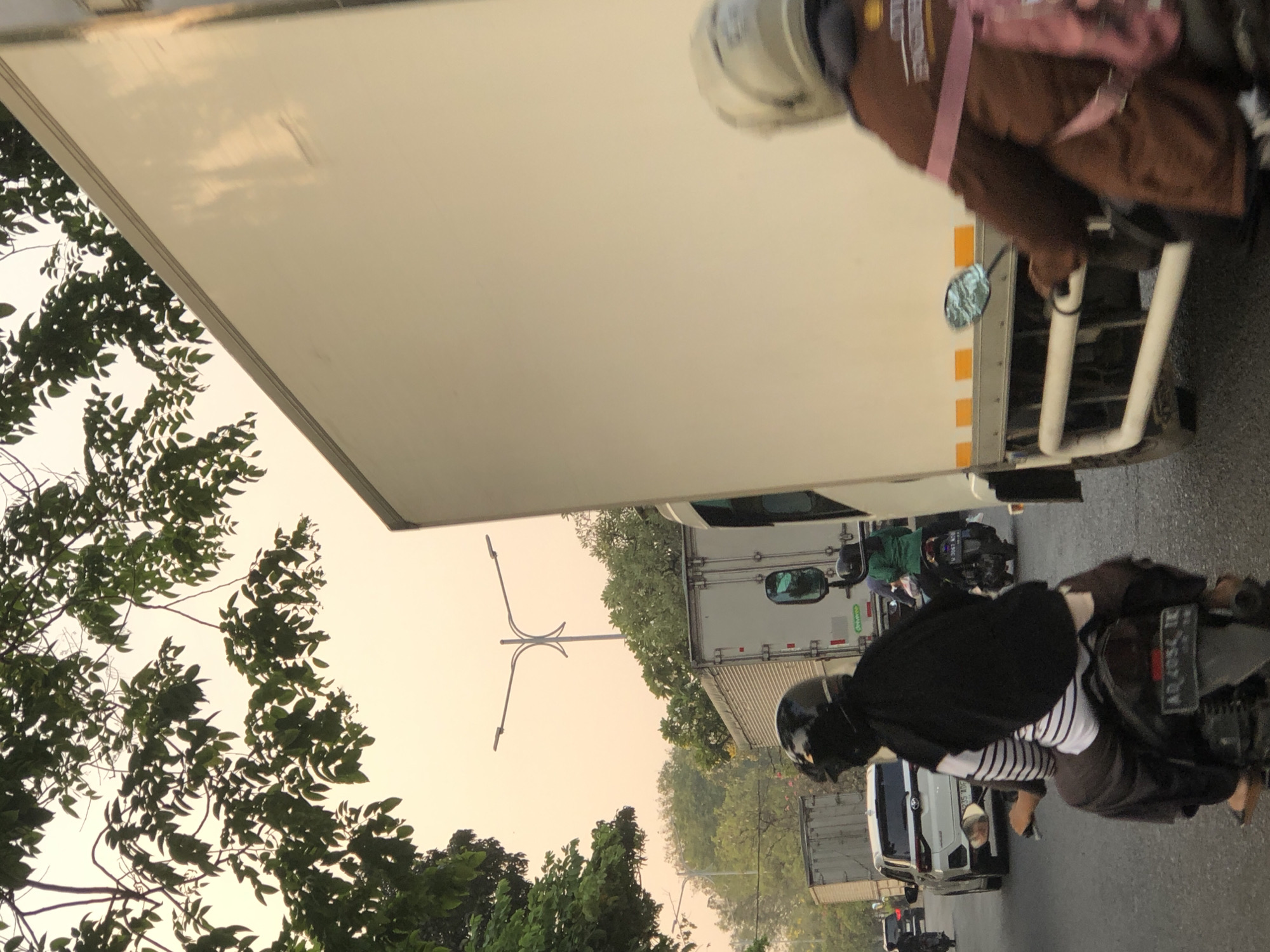 The image depicts a street scene in Sumorame, Indonesia, characterized by heavy traffic and an overcast sky. In the foreground, two individuals on motorcycles are visible. The first, on the left, is wearing a black helmet and a black and white striped shirt, with a black headscarf. They are seated on a motorcycle with license plate "AD 4054 TK". To the right, another person wearing a light-colored helmet and a brown jacket is on a motorcycle, partially obscured by a large white truck. This truck, taking up a significant portion of the right side of the frame, is white with orange reflective stripes near the bottom. Several other vehicles, including a white SUV with license plate "AD 1067 ADF" and other trucks, are present in the background, indicating a busy road. Overhead, tree branches with green leaves extend into the frame, and a distinctive street lamp with multiple arms is visible in the distance against the hazy, pale sky. The overall atmosphere suggests a typical day in an urban environment with moderate traffic congestion.