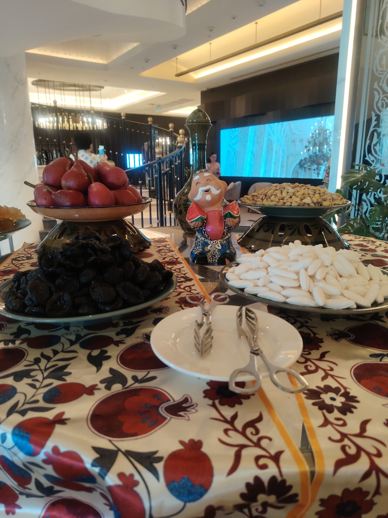 A low-angle, indoor shot shows a table laden with a variety of dried fruits and nuts, arranged in an establishment in Konigil, Uzbekistan. In the foreground, a white plate holds a pair of silver serving tongs. Behind it, a large platter is filled with white, oblong candies. To the left, a dark plate overflows with dried prunes. Above the prunes, a tiered stand displays a pile of red pears. Further to the right, another platter holds a mound of pistachios, and a small, ornamental figure with a red hat stands to the left of the pistachios. The table is draped with a cloth featuring a pattern of red pomegranates and dark leaves. In the background, a large screen displays a blue-toned image of an ornate interior, possibly a palace. A dimly lit staircase with a dark railing is visible in the mid-ground, with indistinct figures moving on it. The overall ambiance suggests a buffet or food display setting in a hotel or restaurant.