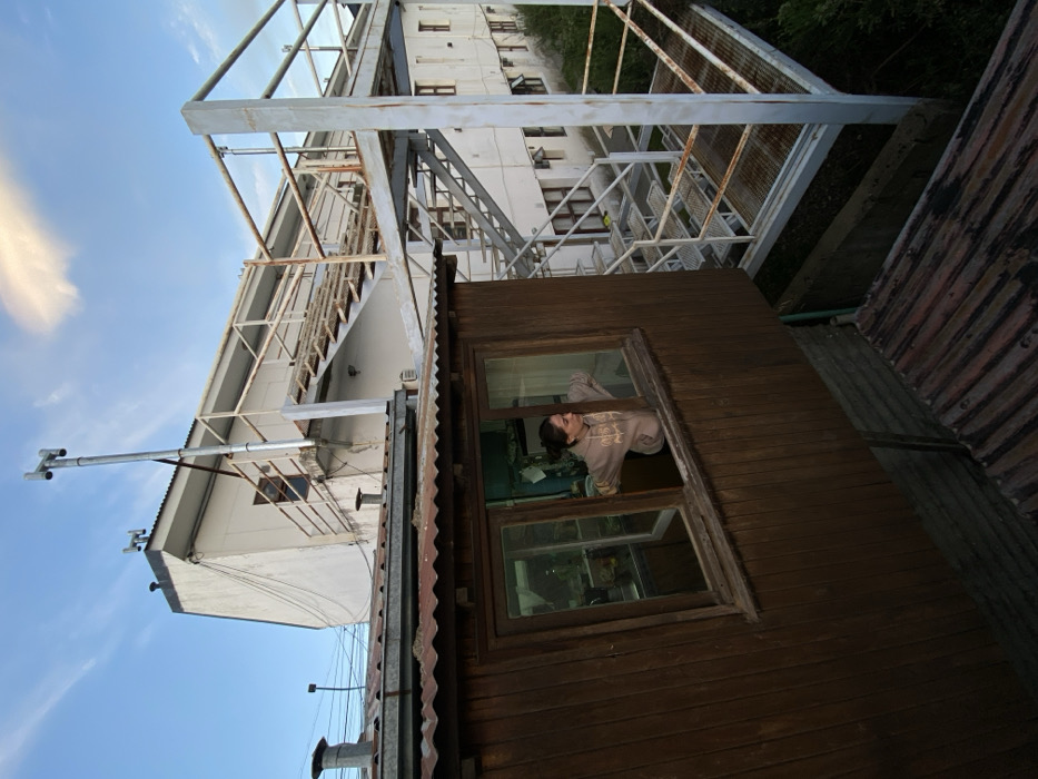 A woman peers out of a window in a dark, wooden shed. The window has two panes and a brown frame. The shed's roof is corrugated metal. A tall, white building with a metal staircase is behind the shed. A metal pole with two small devices extends to the left of the shed. A cloudy blue sky is in the background.