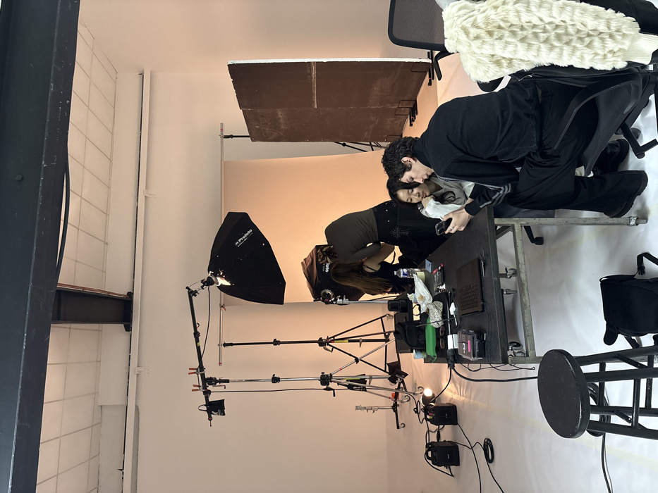 This image captures a behind-the-scenes moment within a photography studio located in Paris, France. The environment features white walls, a white cyclorama floor, and exposed dark ceiling beams.
In the mid-ground, a large black Profoto softbox mounted on a light stand illuminates a white seamless backdrop. Another dark brown rectangular panel stands vertically to the right of the backdrop. Studio lighting equipment, including multiple light stands and power units connected by cables, is positioned on the left side of the frame.
Four individuals are present. On the right, in the foreground, a male with dark curly hair, dressed in a black long-sleeved top and black trousers, is seated in a black office chair. He holds a smartphone and is actively viewing its screen. An Asian female with long dark hair, wearing a light-colored top, sits beside him, also observing the smartphone screen with a slight smile.
Further into the mid-ground, a female stylist, wearing a dark top and trousers, stands with her back partially to the camera. She is engaged in styling the long dark hair of another female subject, who is seated on a low chair and wears a dark, possibly sleeveless, top. A bottle of water is visible near the seated subject.
A black, rectangular rolling table occupies the center-midground, displaying a silver laptop, various small containers, a green pouch, sunglasses, a white paper bag, a small black box with cables, and other production accessories. A dark wooden stool is positioned in the immediate foreground on the left, with a black bag beneath it. A white, textured garment is draped over an office chair on the far right. The scene depicts a preparation phase within an active studio setting.