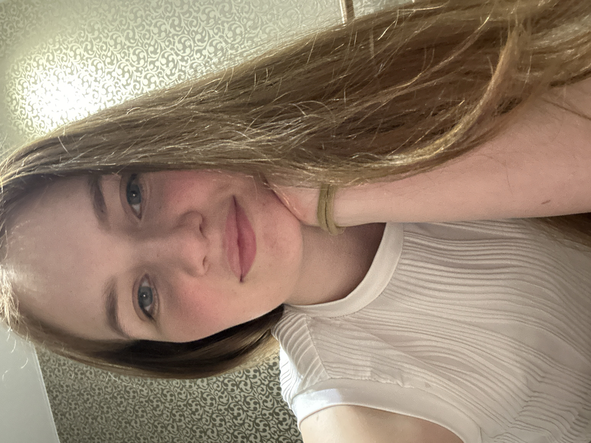 The image shows a selfie of a young woman with long, light brown hair. She has fair skin, light eyes, and a slight smile. She is wearing a white, textured top. Her left arm is bent and her hand is resting on her cheek. The background is a patterned wallpaper, possibly in a hotel room or residential interior. The image is rotated slightly counterclockwise. No information indicates that this image was taken in Chaikovsky, Russia.
