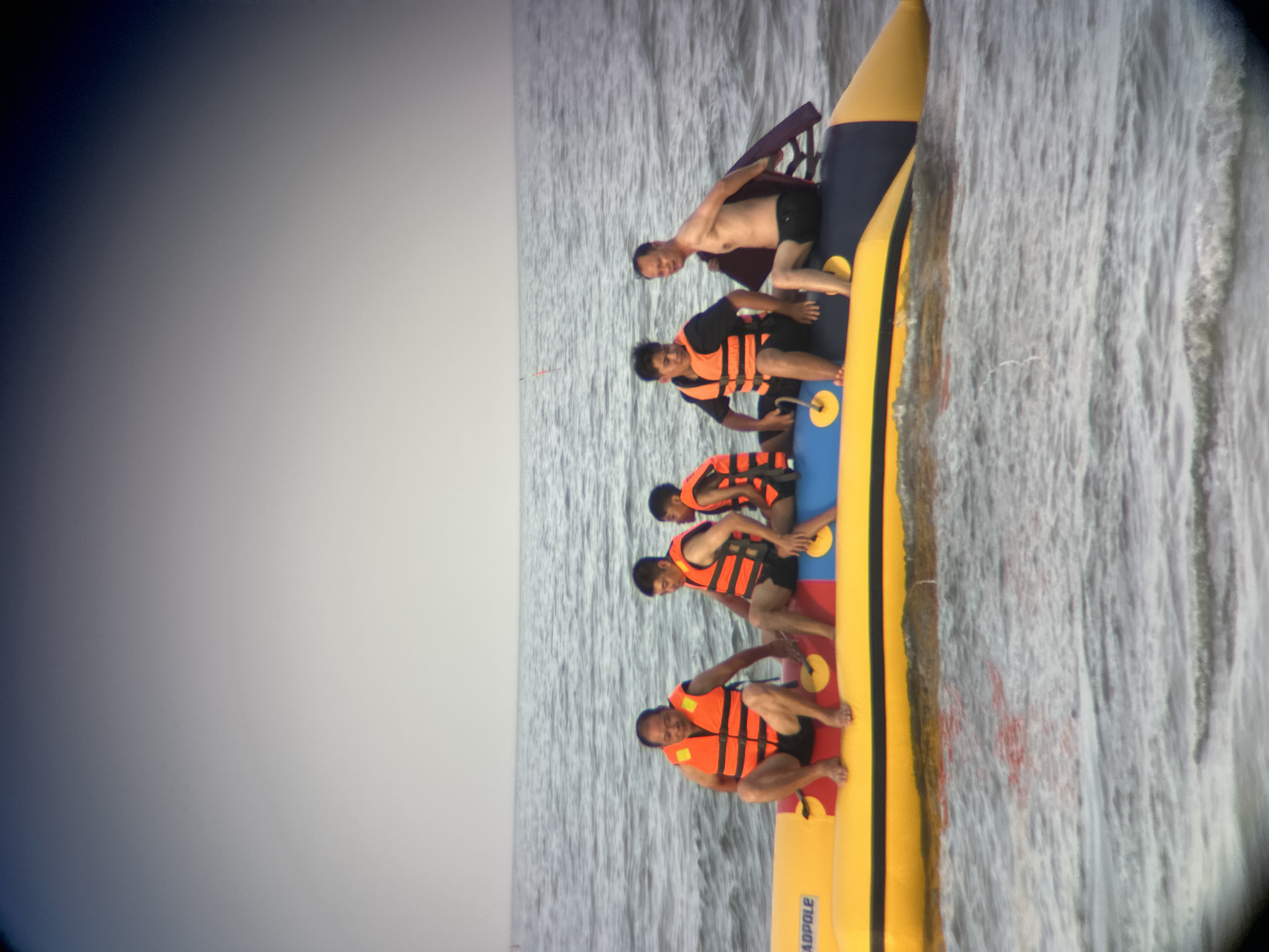 Five individuals are seated on a yellow and blue banana boat in the ocean off the coast of Đà Nẵng, Vietnam. The sky is overcast, and the water is choppy. The individuals, all wearing orange life vests, appear to be enjoying a recreational activity. The banana boat has the word "TADPOLE" visible on its side. The person on the far right is shirtless, wearing black swim trunks, and holding a dark-colored object. The person to their left is wearing a black t-shirt and an orange life vest. The person in the middle is also wearing a black shirt and an orange life vest. The person to the left of them is wearing an orange life vest and a black t-shirt. The person on the far left is wearing an orange life vest and black shorts. The image captures a moment of leisure and water sports in a coastal environment.