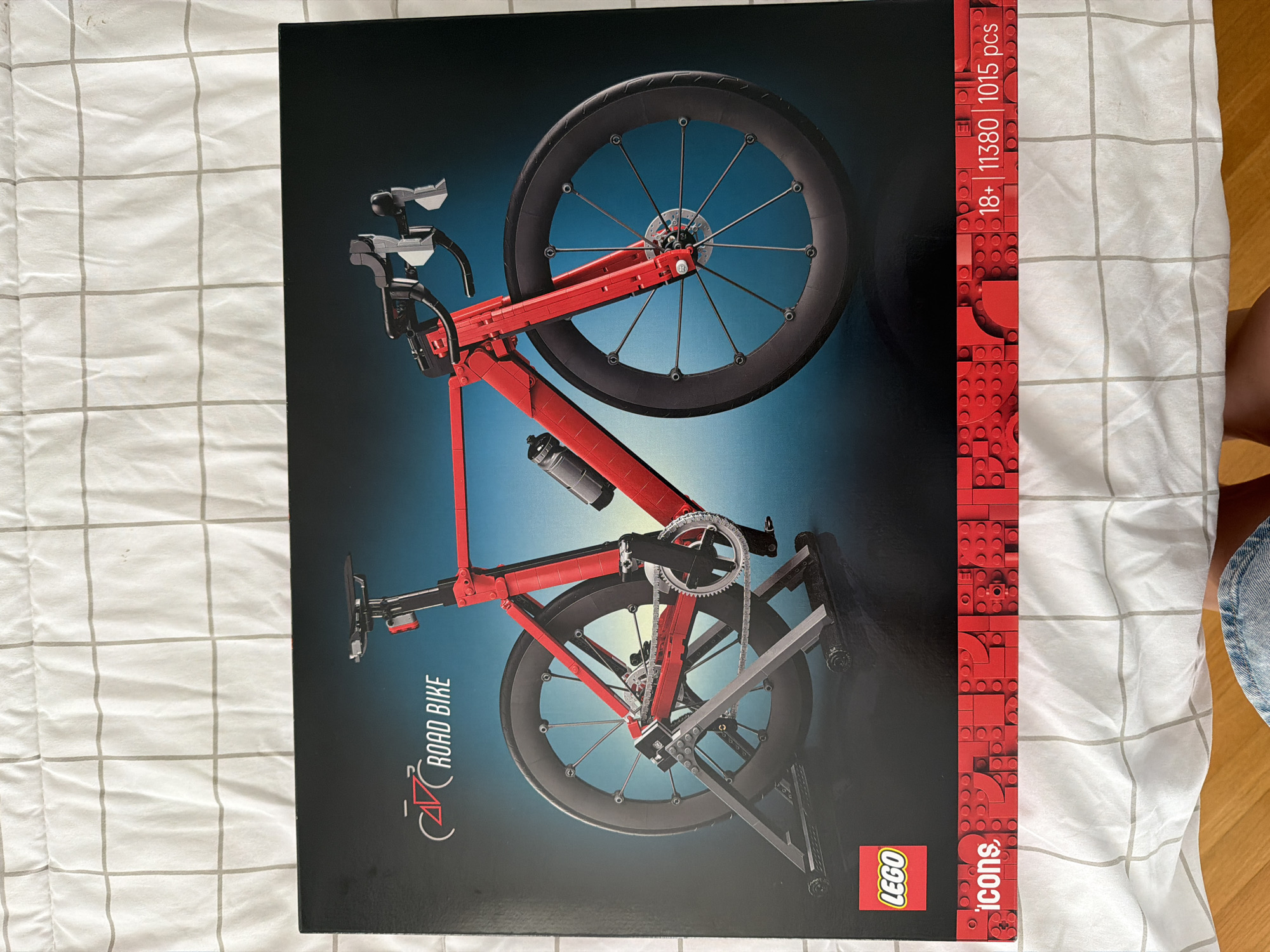 The image shows the front of a LEGO set box, featuring a detailed illustration of a red road bike with black wheels and accessories, presented on a dark blue background. The LEGO logo is visible in the bottom left corner, alongside the "icons" branding. Along the bottom edge of the box, text indicates "18+ | 11380 | 1015 pcs". The box itself is placed on a white, checkered surface, likely a bedspread, with a glimpse of wooden flooring and a person's legs at the bottom edge. There is no indication of the city of Sofia, Bulgaria in the image.