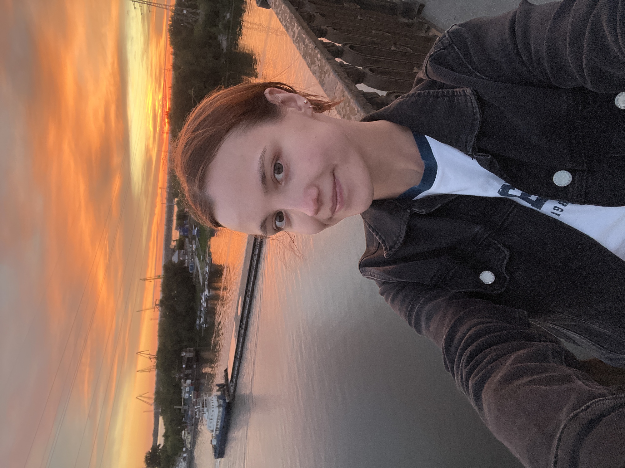 The media file is a selfie taken by a young woman standing on a bridge at sunset. She is wearing a dark denim jacket over a white t-shirt with blue trim. The background shows a vibrant sunset with shades of orange and yellow illuminating the sky, reflecting on a wide river. Several barges and boats are visible on the water, along with industrial cranes and trees lining the opposite bank. The scene appears to be a port or industrial area. The woman is smiling gently at the camera, her expression conveying a sense of peace and contentment. The time of day is clearly sunset, indicated by the dramatic colors in the sky. The location cues suggest an industrial riverside area in Samara, Russia, possibly during a warm evening. There is no visible text in the image.