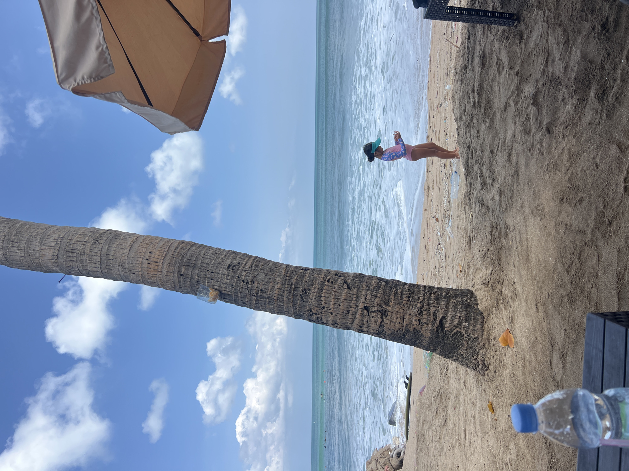 A young girl in a pink rash guard and a blue visor stands on a sandy beach, looking at her phone. The ocean waves gently roll onto the shore, and the sky is a clear blue with scattered white clouds. A large palm tree trunk is prominently in the foreground, leaning to the left. To the left of the palm tree, a tan umbrella is partially visible. In the background, the ocean stretches to the horizon with a few small figures visible in the water, possibly swimmers or surfers. The sand appears somewhat littered with debris. A plastic water bottle with a blue cap is in the lower right corner. The overall scene suggests a sunny day at the beach.