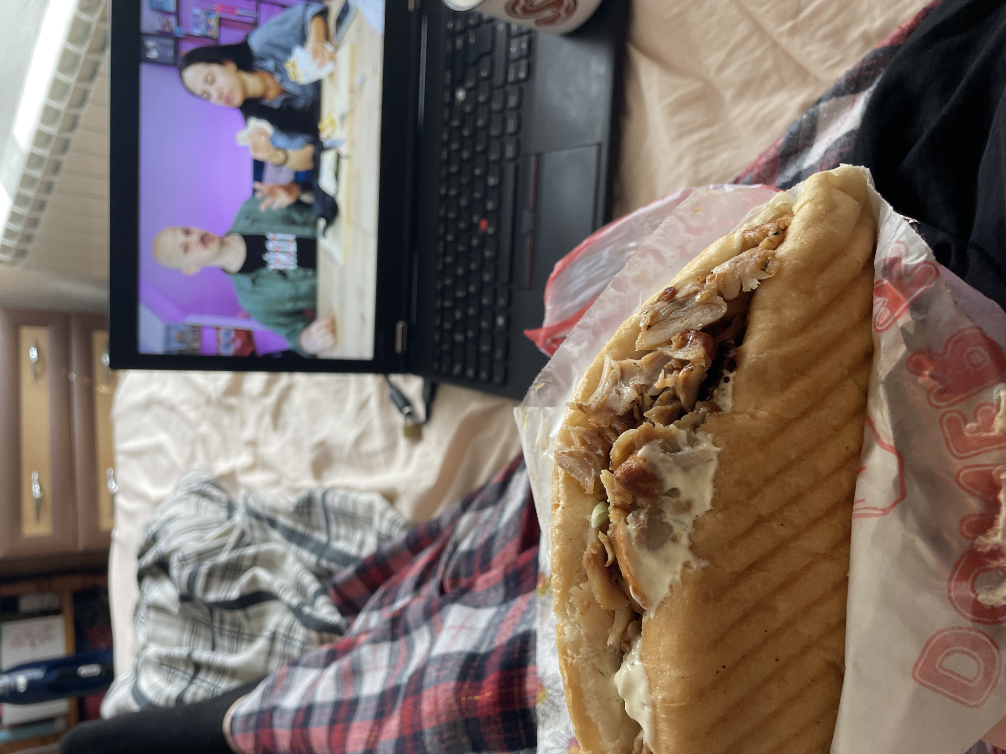 The image shows a close-up of a Döner kebab, partially wrapped in branded paper, alongside a laptop displaying a video. The laptop rests on a bed with a patterned blanket. A mug is visible behind the laptop. The location is presumed to be indoors. There is no definitive evidence in the image to confirm it is in Zlatograd, Bulgaria.