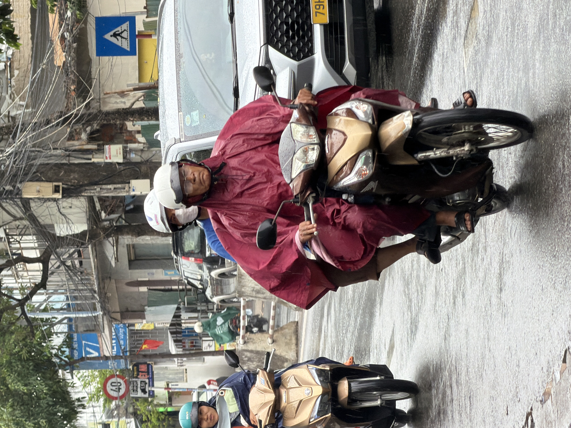 On a wet, overcast day in Nha Trang, Vietnam, a man and a woman are riding a scooter through city traffic. The man, wearing glasses and a red poncho, is in the driver's seat, with the woman behind him, also wearing a poncho. They are both wearing helmets. The road is wet from rain, with puddles visible. In the background, other vehicles, including cars and scooters, can be seen, as well as buildings and street signs. The image captures a typical scene of urban life in Vietnam during the rainy season.