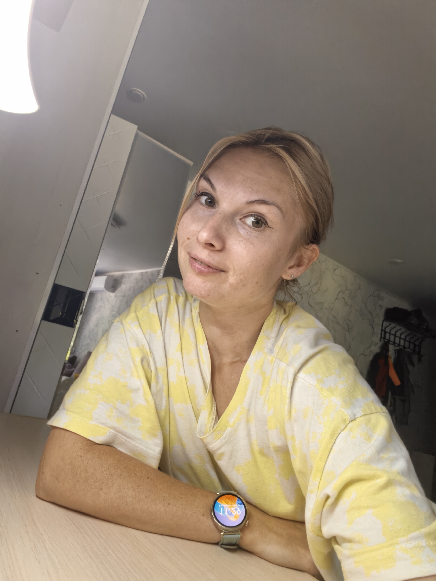A young Caucasian woman with blonde hair, fair skin, and brown eyes is looking into the camera. She is wearing a yellow and white tie-dye t-shirt and a smartwatch on her left wrist. She is sitting at a light-colored wooden desk in what appears to be an indoor setting. The background shows a wall with a marble pattern and some clothing hanging on a rack. The lighting suggests an indoor environment, possibly a home or a small room. The image does not provide any specific information to link it to the city of Sterlitamak, Russia.