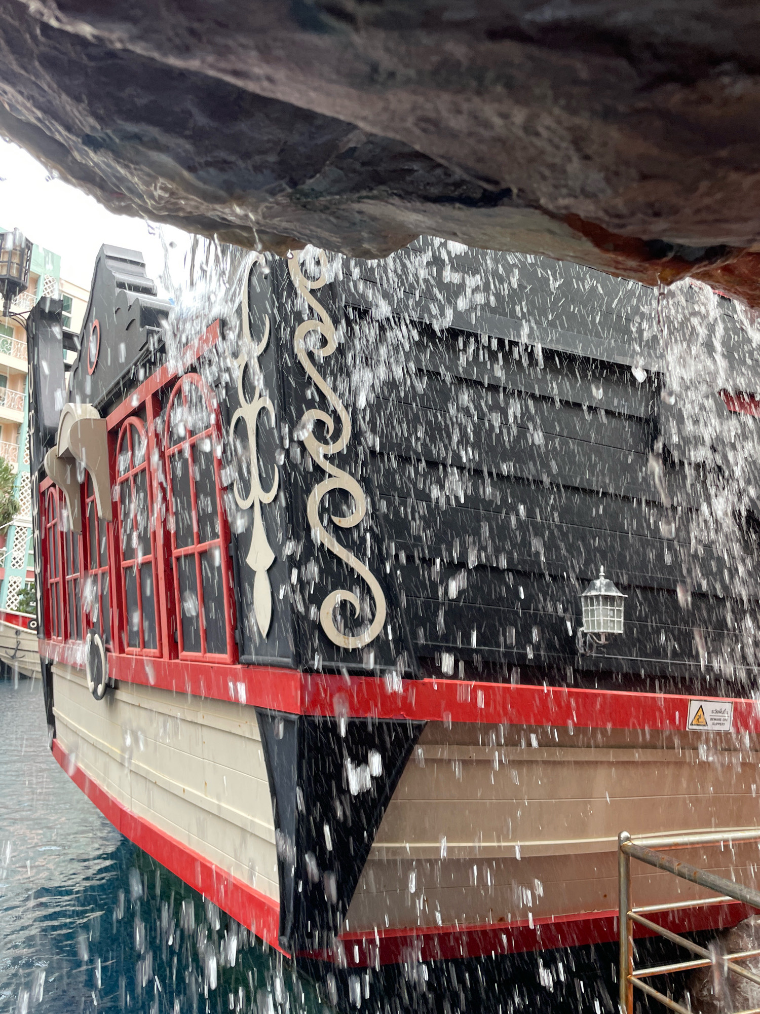 This image shows a close-up view of a themed pirate ship structure, possibly part of an attraction or restaurant, situated in the water. The ship is painted black with red trim and features decorative white swirls on its side. Several red-framed windows are visible along its length. Water is cascading down from an overhead rocky overhang, partially obscuring the ship and creating a dynamic, watery effect. In the background, there are buildings with pastel-colored facades, suggesting an urban or resort setting. The water around the ship is a deep blue. A metal railing is visible in the lower right corner. The overall scene suggests a recreational area within Pattaya City, Thailand, likely designed for entertainment.