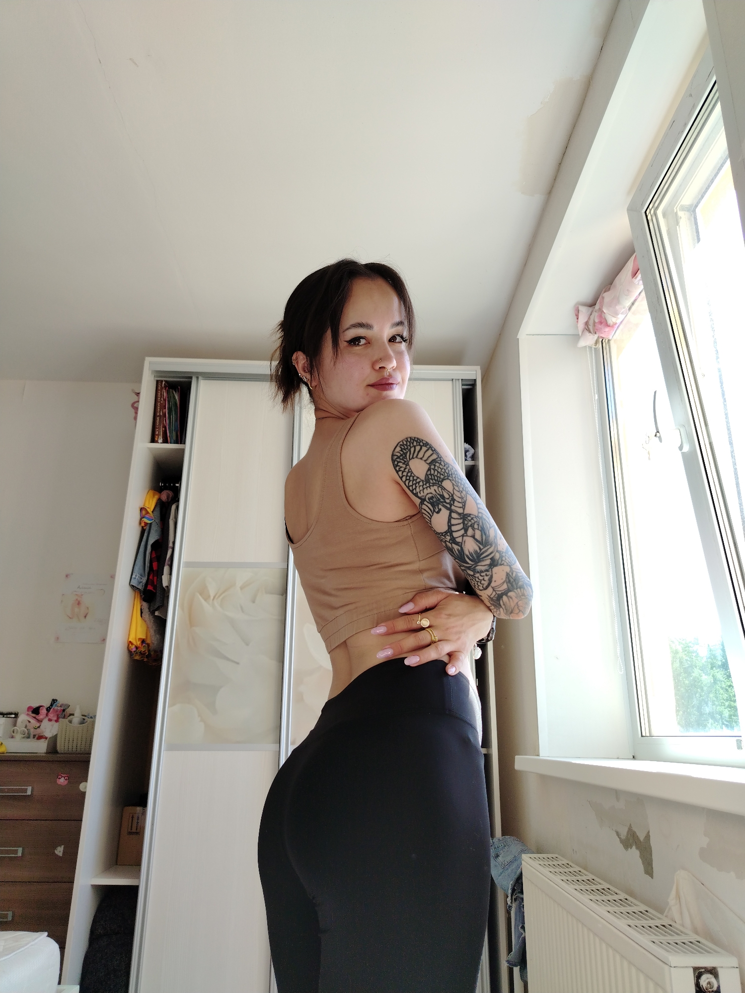 A young woman stands indoors, turning to look over her shoulder at the camera. She wears a tan tank top and black leggings. Her right arm features a detailed black tattoo sleeve. The room appears to be a bedroom, with a large white wardrobe against one wall and a window showing bright daylight. The walls show signs of peeling paint. There is a radiator under the window. The location in the city of Noviy, Russia is not discernible from this image.