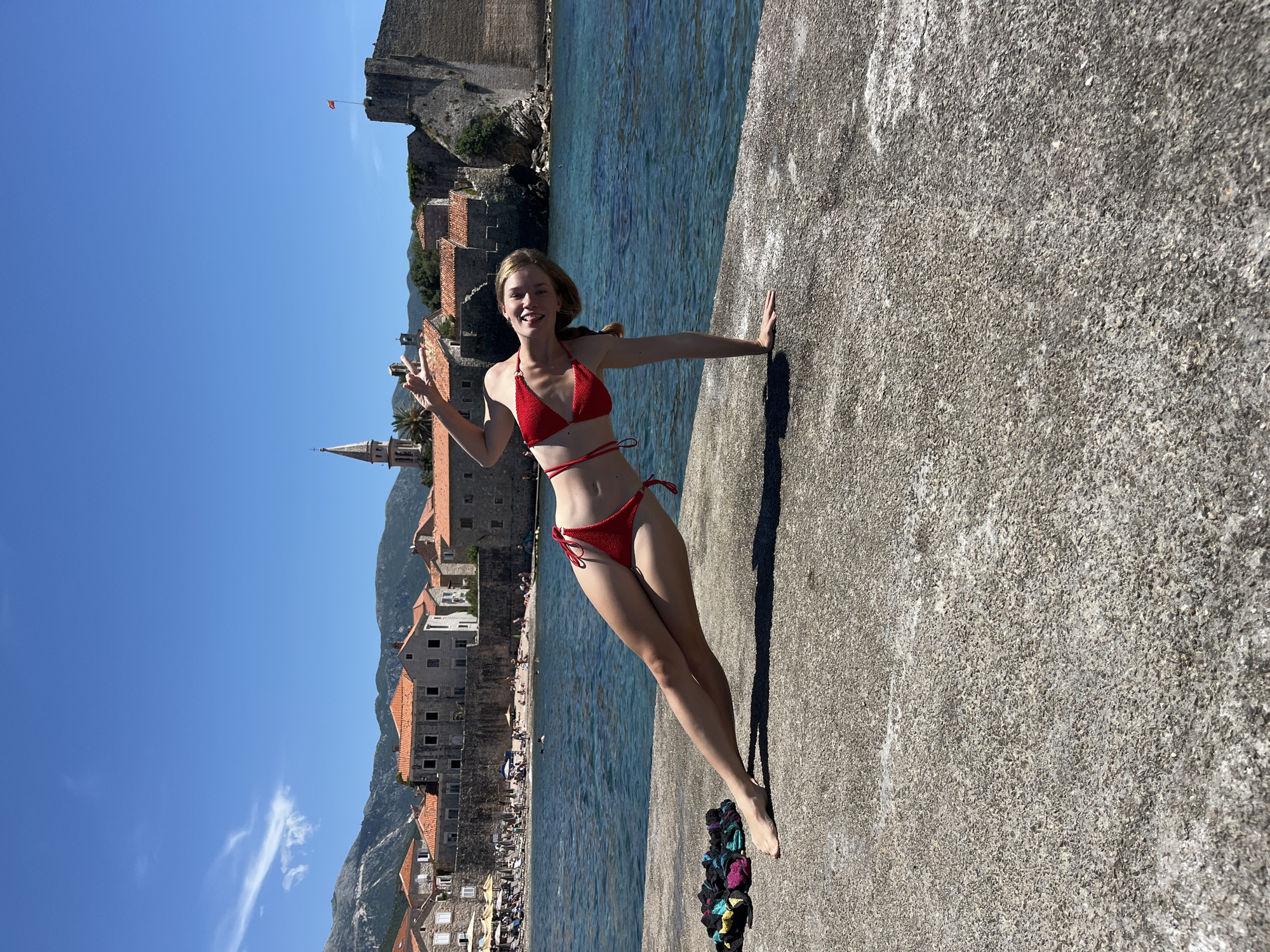A young woman in a red bikini is performing a yoga pose on a concrete pier overlooking the Adriatic Sea. Behind her, the old town of Budva, Montenegro, is visible, with its stone buildings, red-tiled roofs, a church spire, and a fortified castle. The water is a deep blue, and the sky is clear. There are a few people on the beach in the distance, and a small pile of colorful clothing lies on the pier to the woman's left.