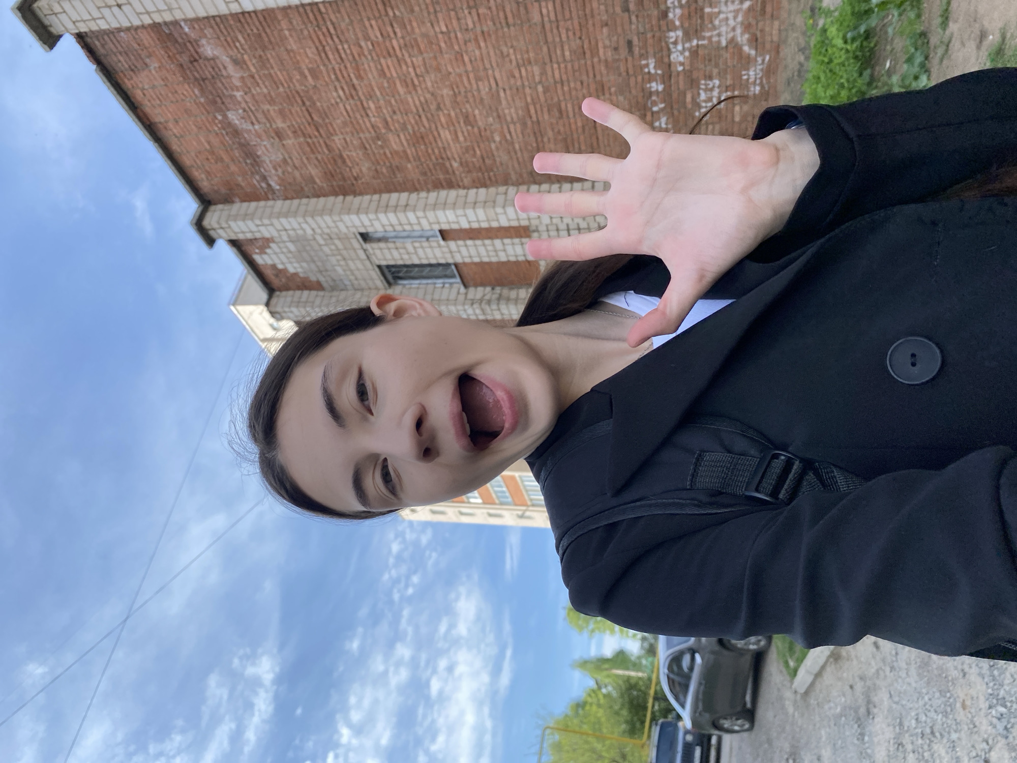A young woman poses for a selfie, contorting her face with her mouth open wide and raising her right hand towards the camera. She wears a black coat over a white shirt and a backpack. The background shows a brick building and a cloudy sky. A car is parked along the sidewalk. The picture was likely taken in a city in Russia.