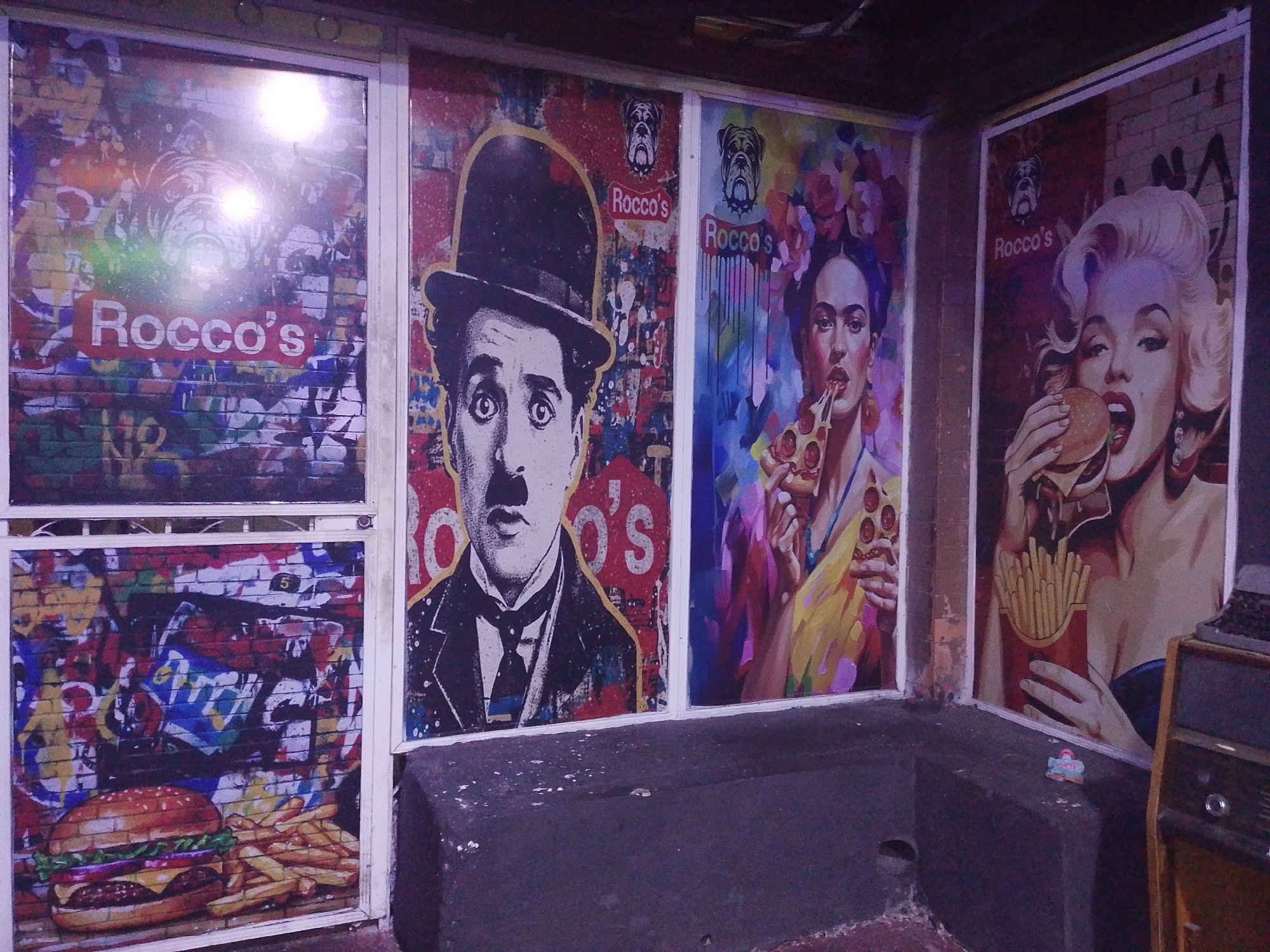 The photograph captures a street scene in Tinaquillo, Venezuela, featuring the exterior of a business. The business's exterior wall is adorned with four framed posters. From left to right, the first poster depicts a hamburger and fries against a graffiti-style background. The second poster features Charlie Chaplin. The third poster showcases Frida Kahlo eating pizza. The final poster displays Marilyn Monroe eating a burger and holding fries. Each poster contains the restaurant's name, "Rocco's", above a bulldog icon. A dark concrete ledge runs along the base of the wall beneath the posters. An old radio sits on the ledge to the right of the posters. The lighting is dim, suggesting the photo was taken indoors or in an area with limited light. The setting appears to be an urban environment with a grunge aesthetic.