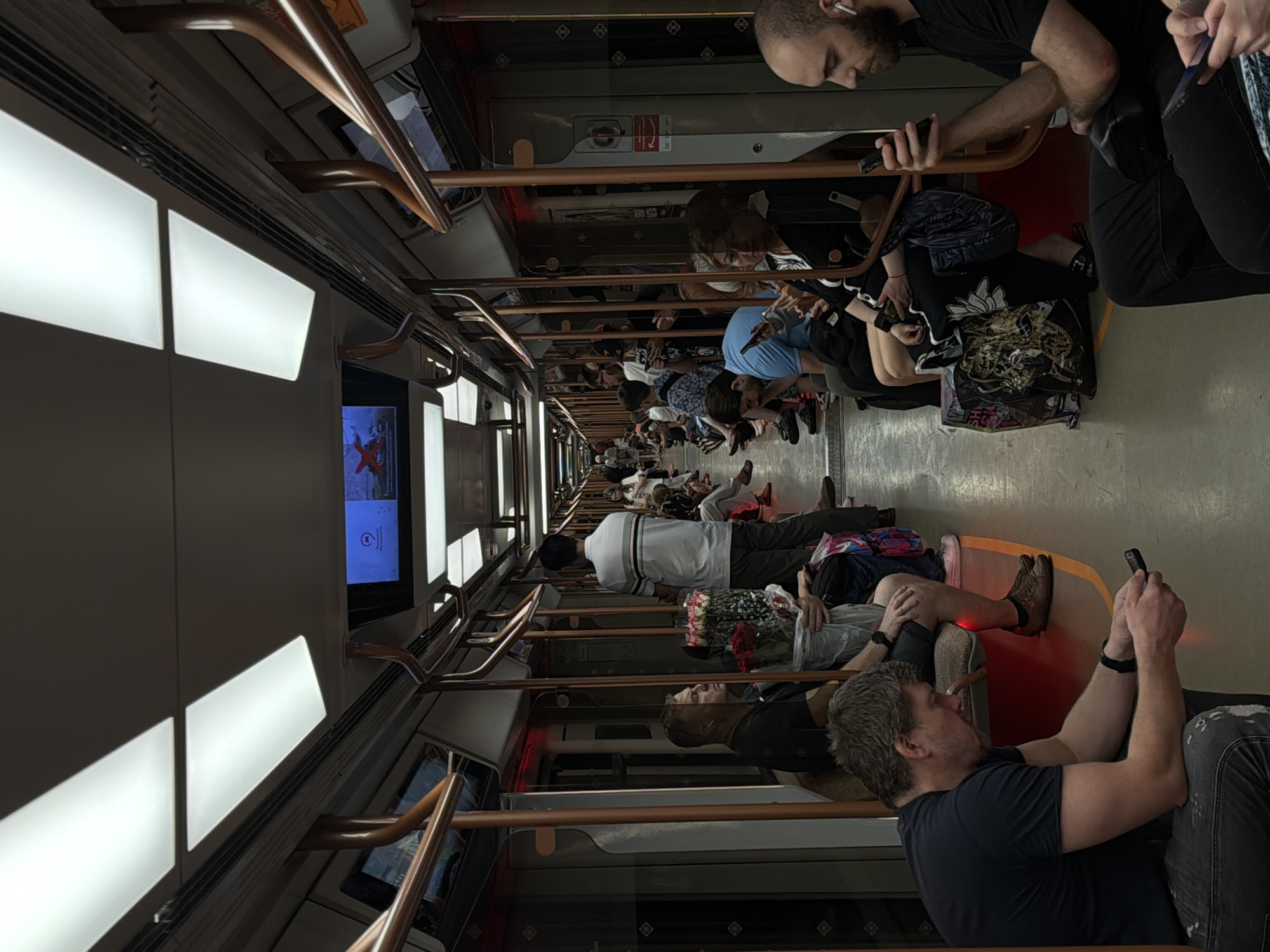 A public transportation scene inside a Moscow Metro train car in Russia shows a densely packed passenger environment. Many passengers are seated, while others stand, all holding onto the copper-colored handrails. Most of the passengers are engaged with their mobile phones. Large light panels illuminate the car's interior. A digital screen displays information on the train's route. The overall atmosphere indicates a typical rush-hour commute or high-traffic time on the Moscow Metro.