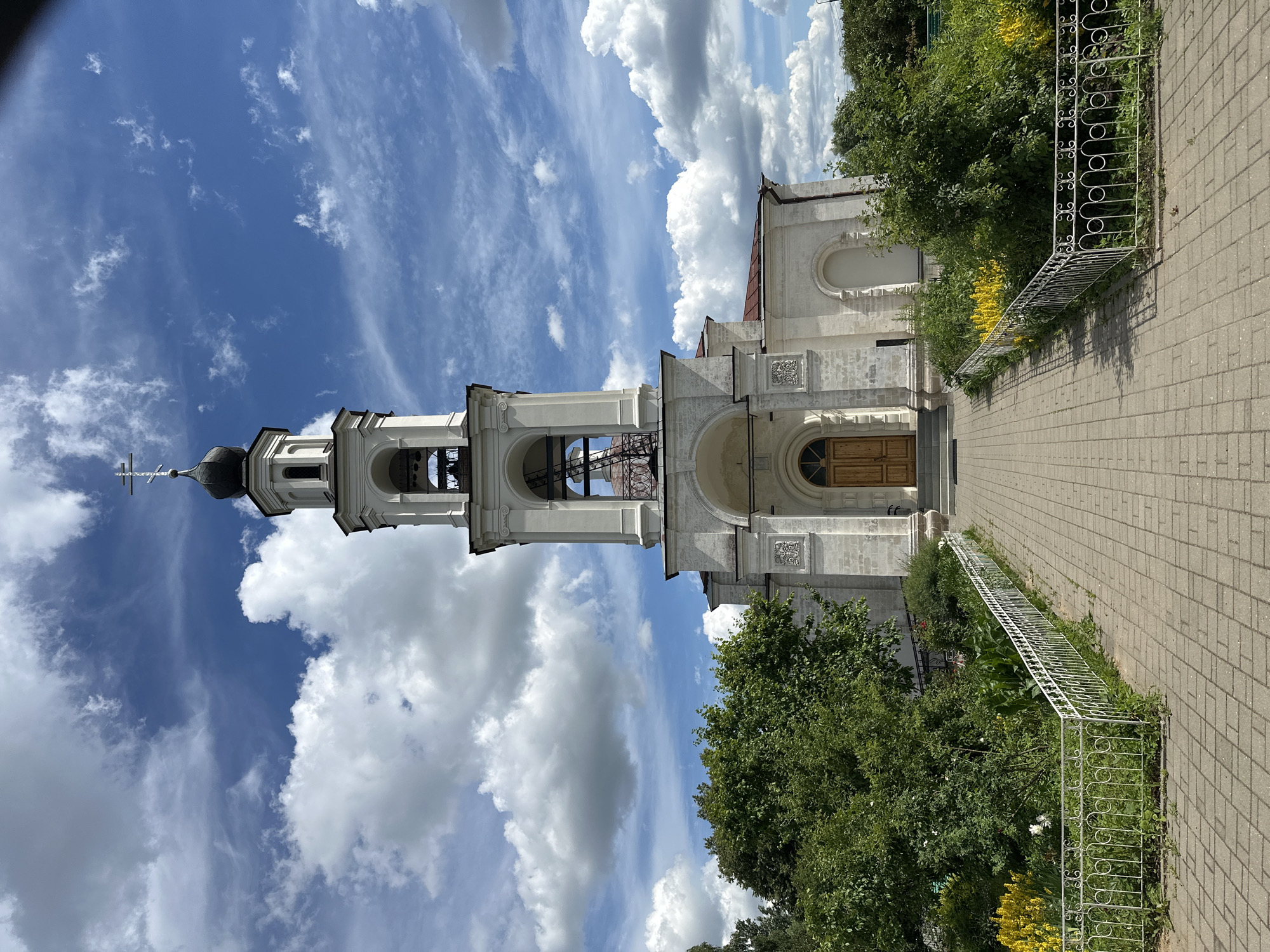 The image captures a bright, sunny day at a religious complex in Ерино, Russia. The primary subjects are a church bell tower and a section of the church building, both constructed from light-colored stone. The bell tower is tall and slender, topped with a dark, domed roof and a cross. It features multiple arched openings where bells would typically hang, and an ornate cross at its apex. Adjacent to the bell tower is the main church structure, also built of stone, with arched windows and decorative architectural details. A wooden door is visible on the church facade, suggesting an entrance.
The scene is set outdoors, with a clear blue sky dominating the upper portion of the image, dotted with fluffy white clouds. Lush green trees and vibrant yellow flowers flank the path leading towards the church. A decorative white metal fence runs along the edge of the path, separating it from the garden area. The ground is paved with light-colored bricks, forming a walkway.
The overall mood is peaceful and serene, characteristic of a quiet day at a place of worship. The bright sunlight and clear sky indicate it is likely midday or early afternoon. There is no visible text in the image.