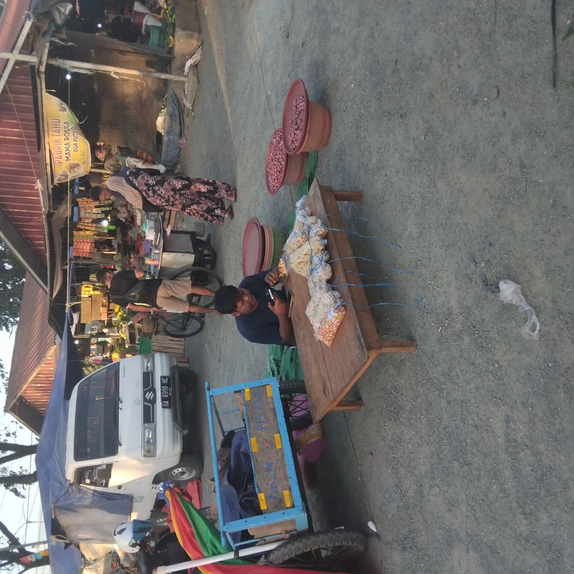The image depicts an outdoor market scene in Pelaihari, Indonesia. In the foreground, a man sits at a wooden table displaying bags of snacks. He is looking at his phone and holding a small object in his hand. The table is set on a gravelly ground, with a blue cart to the left and two large baskets of onions to the right. In the background, a white truck is parked, and market stalls with various goods are visible. People can be seen walking and interacting within the market. The overall environment suggests a bustling local marketplace.