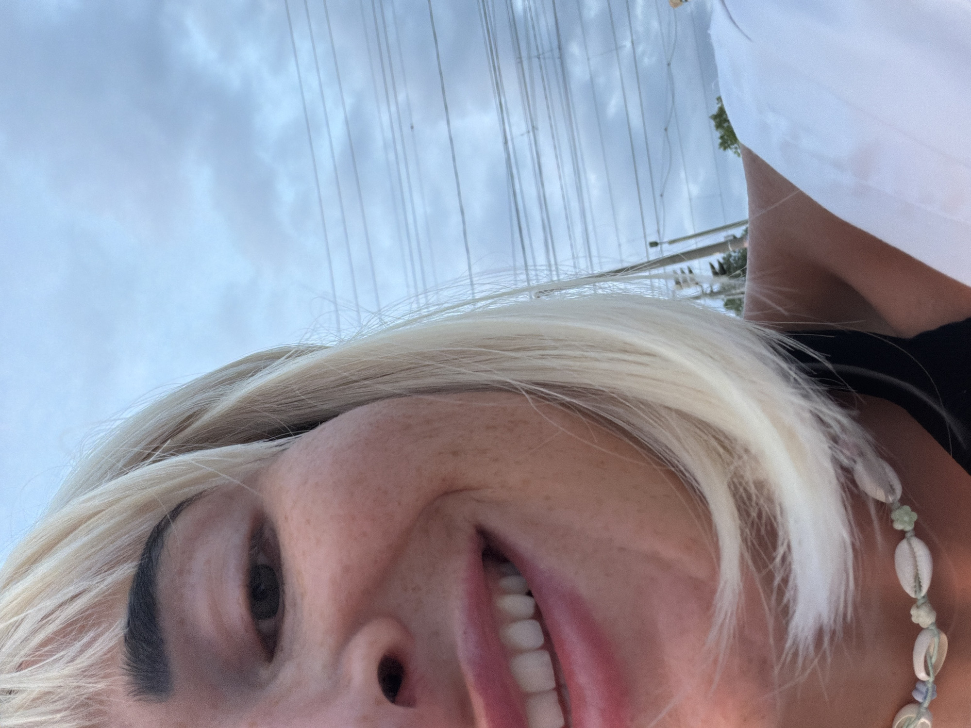 A half-profile, close-up selfie of a woman with blonde hair and fair skin, her left eye and mouth visible. She is smiling, revealing her teeth. The background shows a cloudy sky with power lines, suggesting an outdoor urban or suburban setting. The woman is wearing a necklace made of shells and small flower-shaped beads. The scene appears to be located in the city of Sochi, Russia.