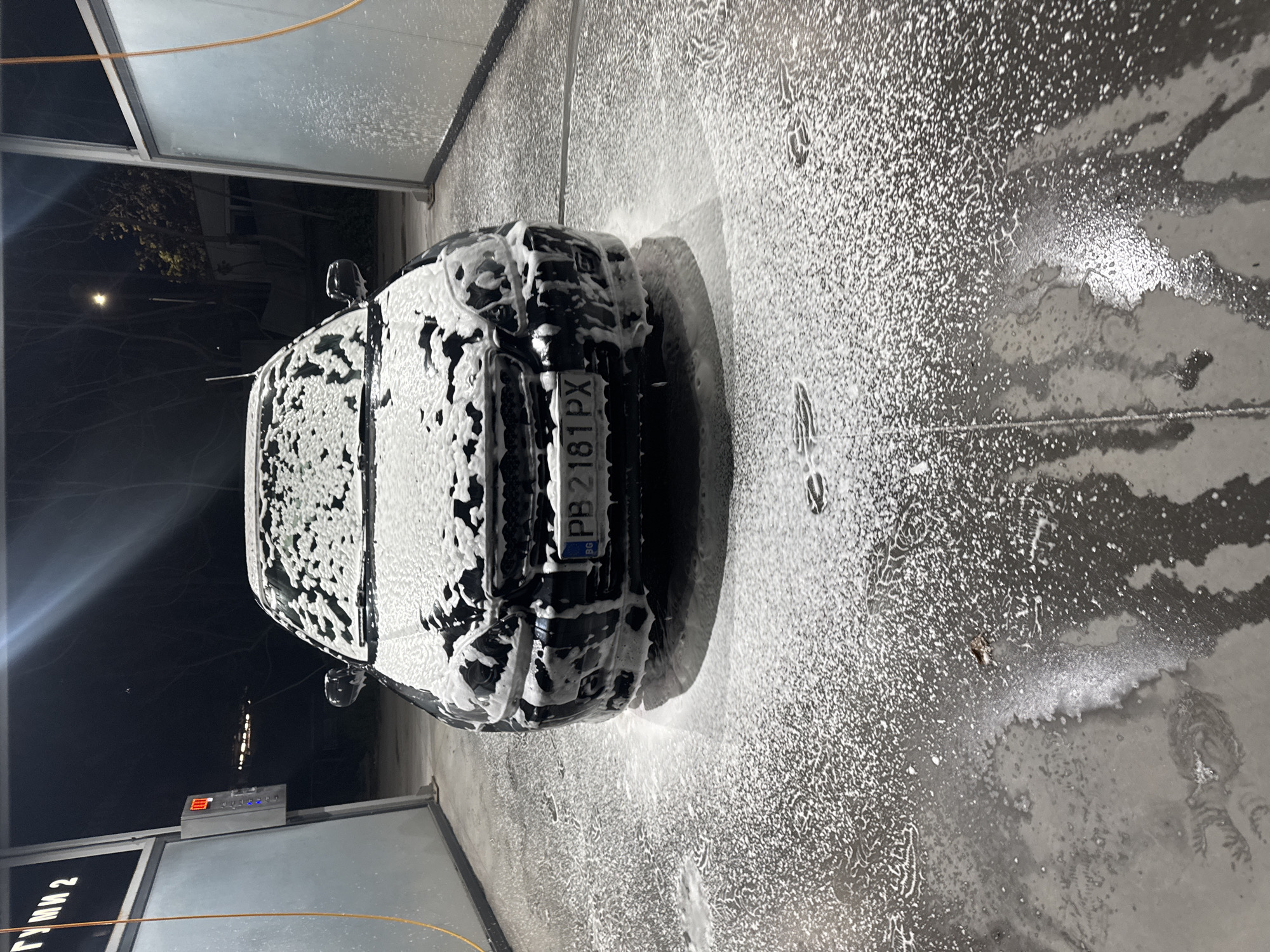 An overhead, vertically oriented photograph, rotated 90 degrees clockwise, captures a dark-colored car completely covered in white soap foam within a car wash bay at night. The car is positioned with its front facing towards the bottom of the image (or right side in the rotated view). Its license plate, "PB 2181 PX", is clearly visible on the front bumper, along with the "BG" country code on a blue strip, indicating Bulgaria, specifically Plovdiv (PB).
The car wash bay is illuminated, contrasting with the dark night sky visible outside the structure. The concrete floor of the bay is dark and wet, covered in scattered foam and water, showing reflections of light sources. Several foamy footprints or marks are discernible on the wet floor.
On the left side of the bay, a control panel or payment terminal with illuminated buttons and a digital display showing "E8" is visible. Below it, a sign with partially visible text reads "ГУМИ 2" (GUMI 2), likely indicating a bay number or related service. An orange hose or cable runs across the upper left portion of the image. The general activity depicted is a car being washed using foam, likely during a self-service car wash.