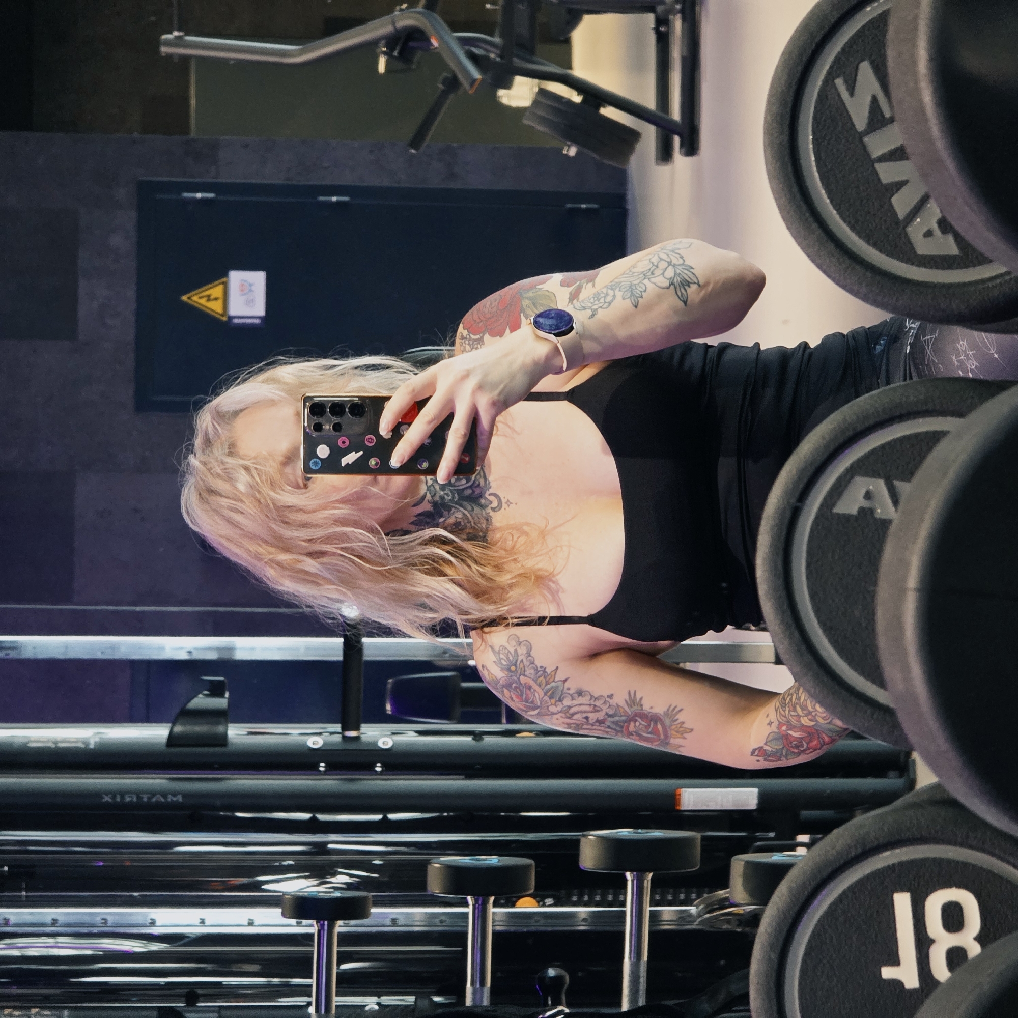 A woman is taking a selfie in front of a mirror at a gym. The gym has weight racks holding weight plates in the background. The woman has blonde hair, tattoos on both arms and chest, and is wearing a black tank top. She is holding a smartphone with a decorated case to take the photo. The brand name "MATRIX" is visible on the equipment. A yellow warning sign is on the wall behind her. The location is Saint Petersburg, Russia.