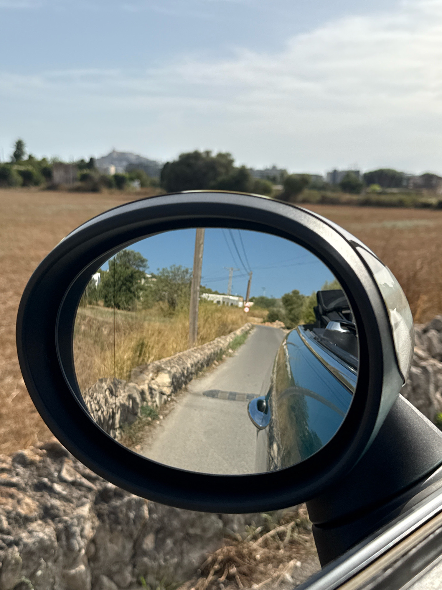 The image shows a reflection in a car's side mirror, displaying a paved road winding through a rural landscape. The road is bordered by a low stone wall and dry, tall grass. In the reflection, trees and white buildings are visible, with a distant hill topped by structures, possibly a town or village. The sky is clear and blue. The exterior of the car, a dark teal convertible, is partially visible in the foreground, with its roof down. The image was captured on a sunny day. The distant hill with structures is consistent with the appearance of Santa Eulària des Riu, Spain, known for its hillside architecture and coastal setting.