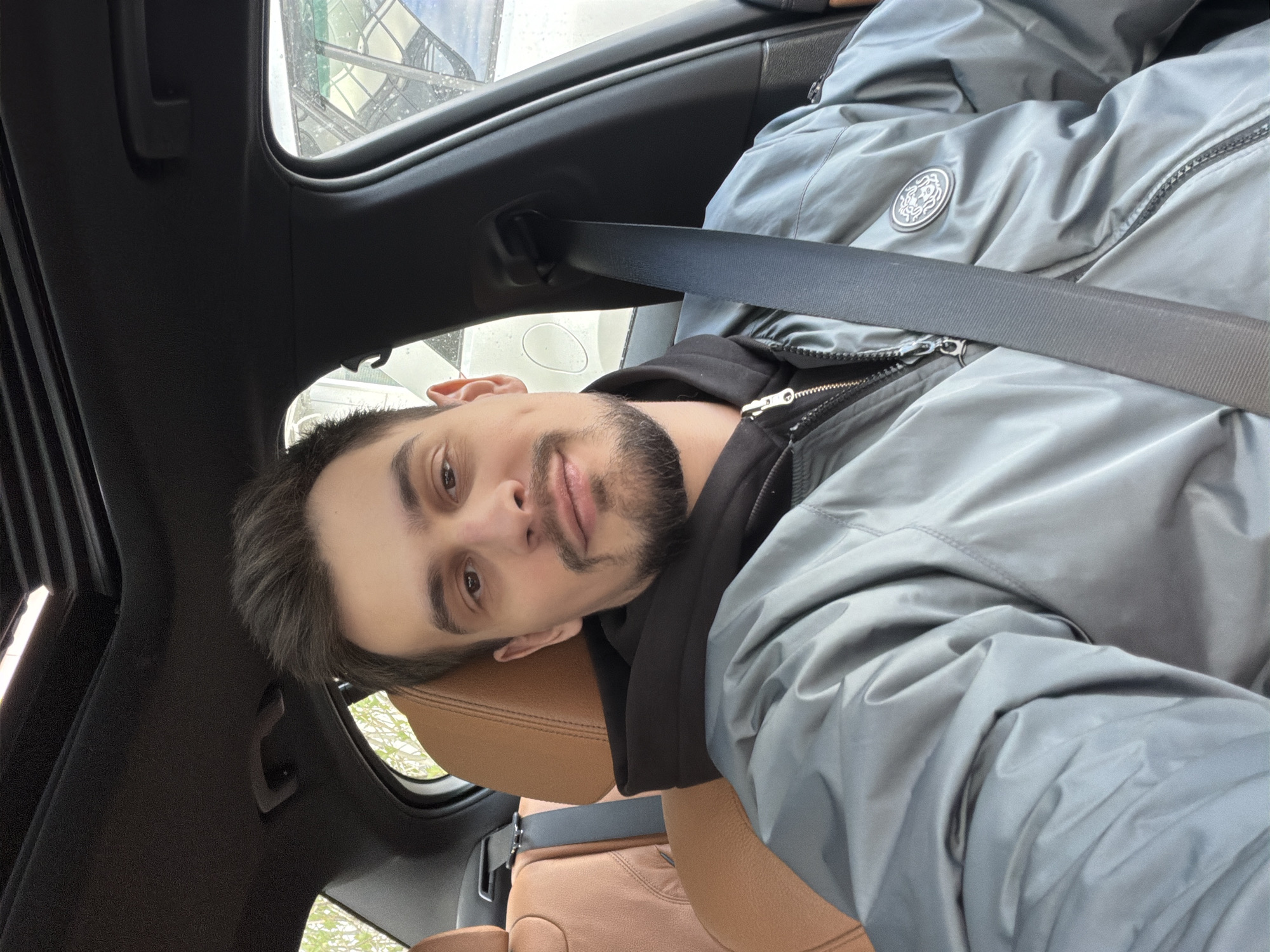 A person is seated in the front passenger seat of a car. The car is black with light brown/tan leather seats. The person is wearing a light gray jacket and a black hoodie, with the seatbelt fastened. The person appears to be male, with short brown hair and a short beard. The photo is taken from an odd angle, with the camera pointing up at the person. The background shows a glimpse of the outside, including what appears to be a building. Without additional information or clearer context, it's not possible to determine if the location is in Krasnoyarsk, Russia.