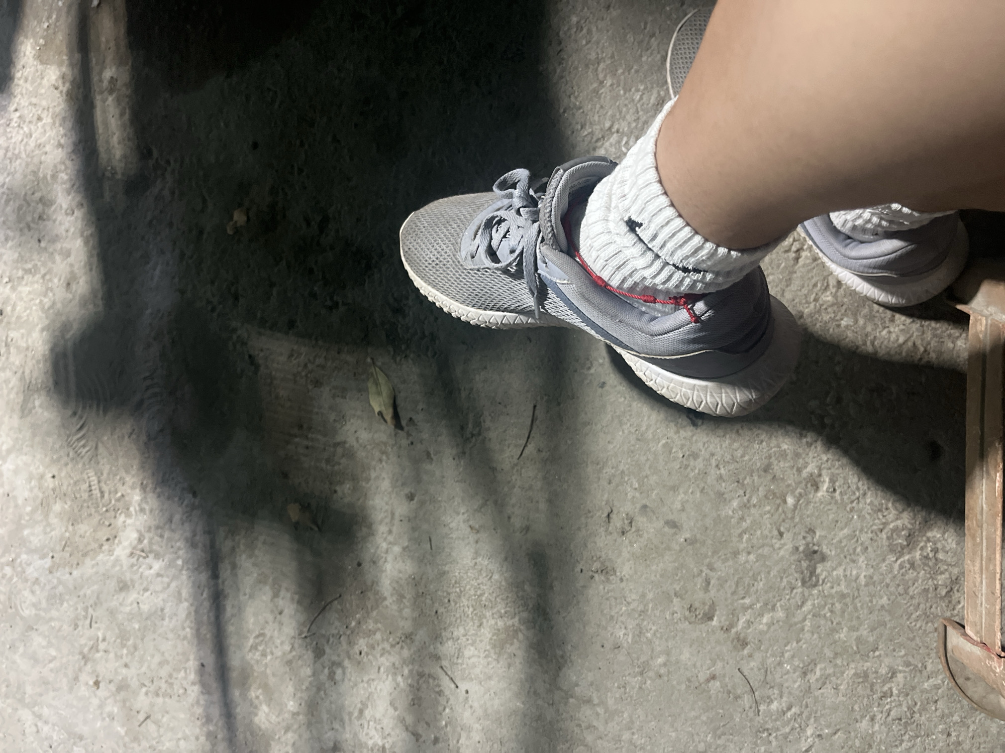 The image provides a high-angle, close-up view of a person's lower leg and foot, clad in an athletic shoe and sock, set against a concrete ground. The lighting is dim, suggesting night or low-light conditions, with strong shadows cast across the rough, grey ground surface.
The central subject is a grey athletic shoe with a mesh upper, white patterned soles, and grey laces. A small red detail is visible near the heel of the shoe. The foot is covered by a white, ribbed crew sock, pulled up around the ankle, with the person's bare lower leg visible above.
The ground is a textured, light to dark grey surface, likely concrete or asphalt, with scattered small debris and dried leaves. Prominent, dark shadows dominate parts of the foreground and background, indicating a singular, angled light source, possibly artificial. On the far right edge of the frame, a segment of a metallic, reddish-brown object, resembling a structural support or furniture leg, is partially visible.
No specific activity is occurring; the foot appears to be stationary. There is no visible text within the image. The scene is located in Santa Ana, Venezuela.