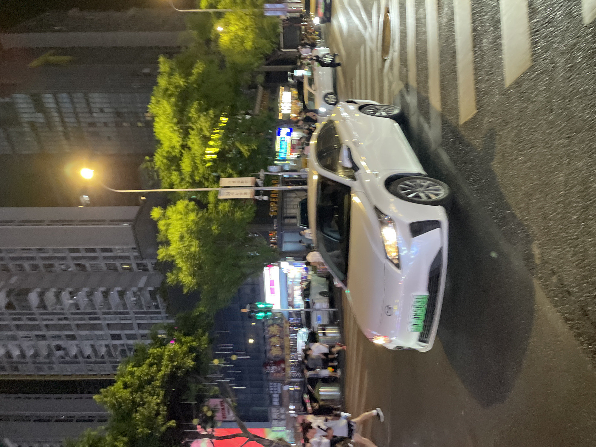 The image depicts a nighttime street scene in Longta, China. A white car is stopped at a crosswalk, its headlights illuminated. The car has a green license plate with the characters "A AM5631". Several pedestrians are visible on the sidewalk, some walking and others standing. In the background, tall buildings and trees line the street. Streetlights and neon signs illuminate the scene, creating a vibrant urban atmosphere. The overall impression is one of a busy city street at night, with activity and movement.
