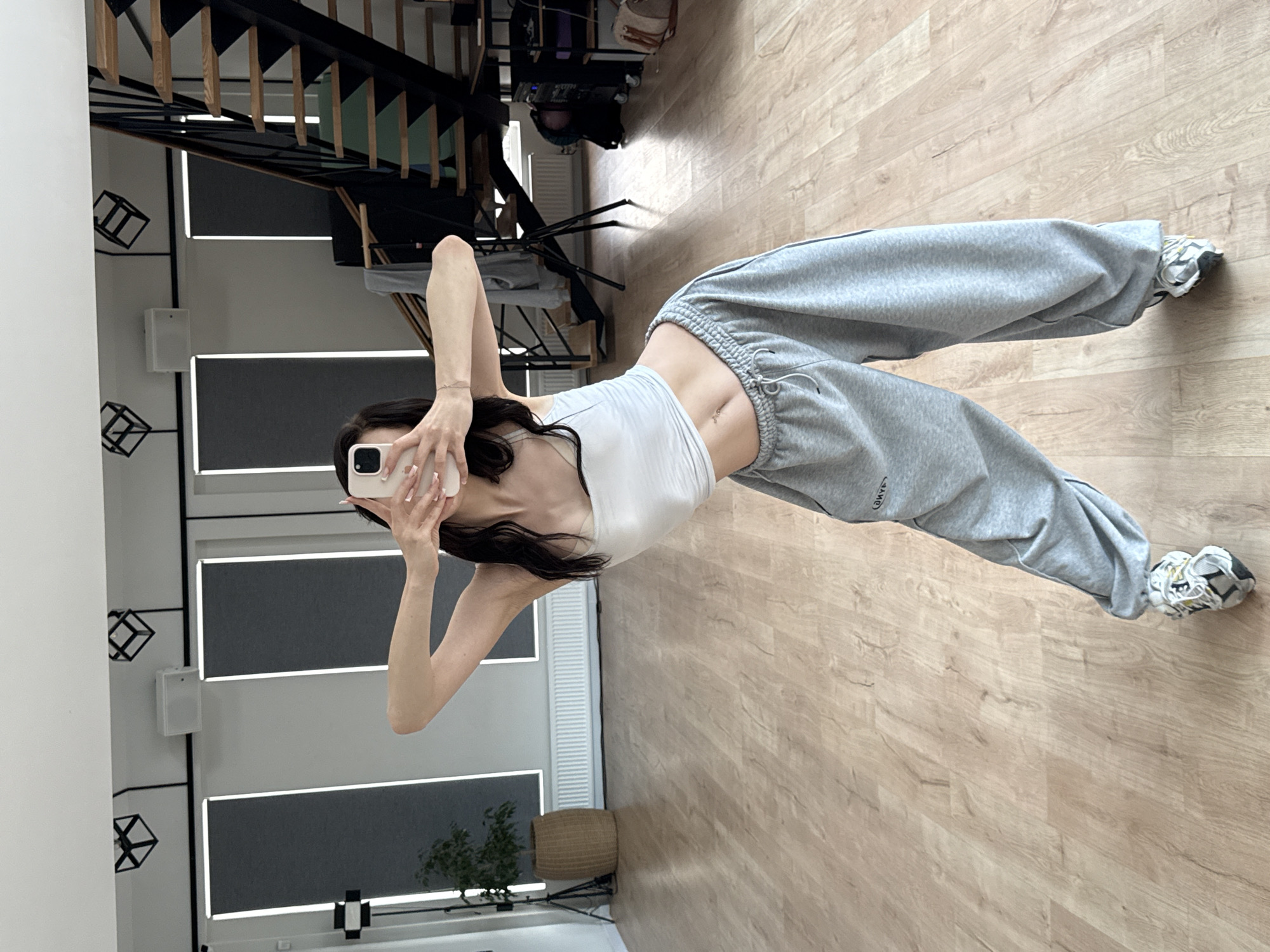 A person is taking a mirror selfie indoors, likely in a dance studio or a room with a wooden floor and a staircase in the background. The person is wearing a white crop top and grey sweatpants, with white and grey athletic shoes. Their pose suggests they might be a dancer, as their body is contorted in a way that shows flexibility. The room appears to be well-lit, possibly with natural light from windows, indicated by the illumination of the wooden floor and the reflection in the mirror. The time of day is not specified but the lighting suggests it's daytime. There's a wicker basket with a plant next to a set of windows with dark blinds. A modern staircase with black metal railings and wooden steps is visible in the reflection. The overall mood seems casual and relaxed, as the person is dressed comfortably and engaged in a common social media activity. No visible text is present in the image.