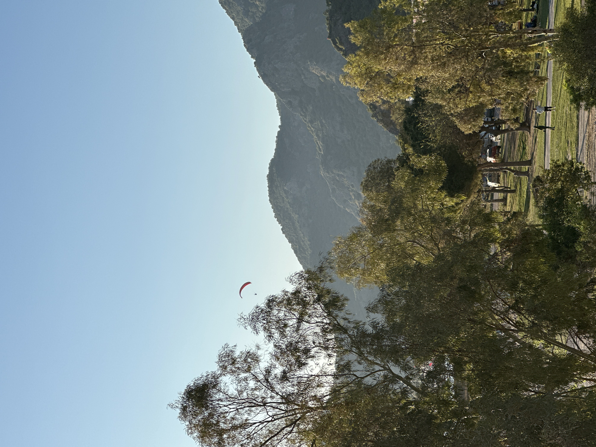 This image depicts a vibrant outdoor scene, likely captured during the golden hour in Fethiye, Turkey, a renowned paragliding destination. The overall scene is dominated by a majestic, rugged mountain range on the left, its slopes covered in dense vegetation, receding into a soft haze.
High in the clear, bright blue sky, a striking red and orange paraglider with a single pilot is visible, soaring gracefully against the vast expanse. This highlights the central activity of paragliding.
In the midground and foreground on the right, numerous trees with rich, leafy canopies, possibly eucalyptus or similar species, are bathed in warm, dappled sunlight. Their branches are silhouetted against the bright sky and illuminated by the sun, indicating a late afternoon or early morning setting. Long shadows stretch across the ground beneath the trees.
On the ground level, beyond the trees, there's a grassy area with a visible paved path or road. Several distant figures (people) can be observed walking or standing, suggesting leisurely activities in a park-like or recreational area. Some vehicles, possibly cars or a caravan, are also subtly visible among the trees or near the path.
The weather is clear and sunny, ideal for outdoor pursuits. The image conveys a sense of adventure, freedom, and the natural beauty of the mountainous landscape. There is no visible text in the image.