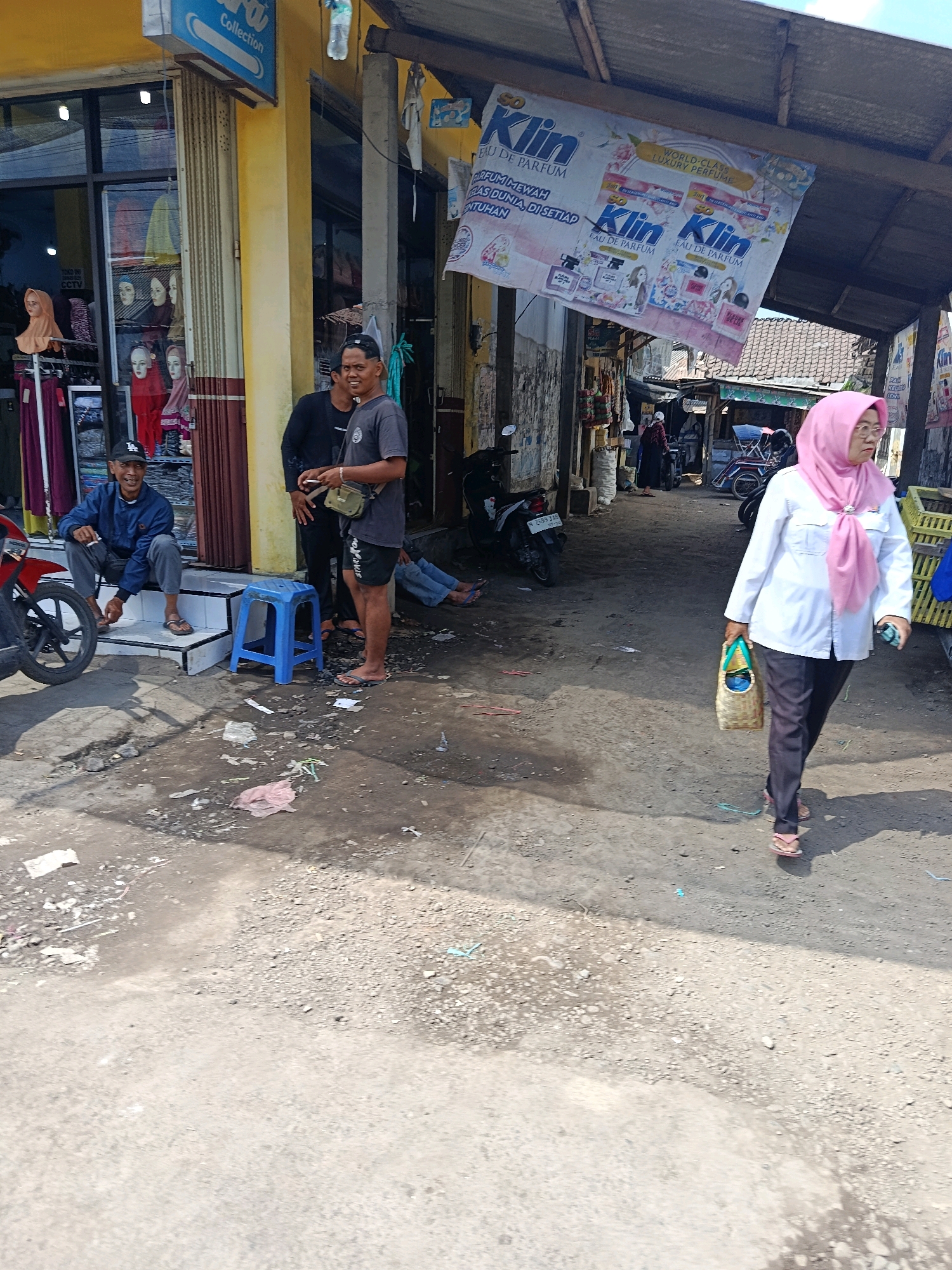 The image depicts a street scene in Jatiroto, Indonesia, likely a market or commercial area. A woman wearing a pink hijab and white shirt is walking towards the right of the frame, carrying a woven bag. In the background, two men are standing and conversing near a storefront. One man is wearing a black shirt and the other a grey t-shirt. A blue plastic stool is placed on the ground near them. To the left, a man in a blue jacket and cap is sitting on steps. Various motorcycles are parked along the street. Overhead, a banner advertising "Klin Eau de Parfum" is visible. The ground is unpaved, with scattered debris and some puddles. The overall environment suggests a bustling, outdoor commercial setting in a town.