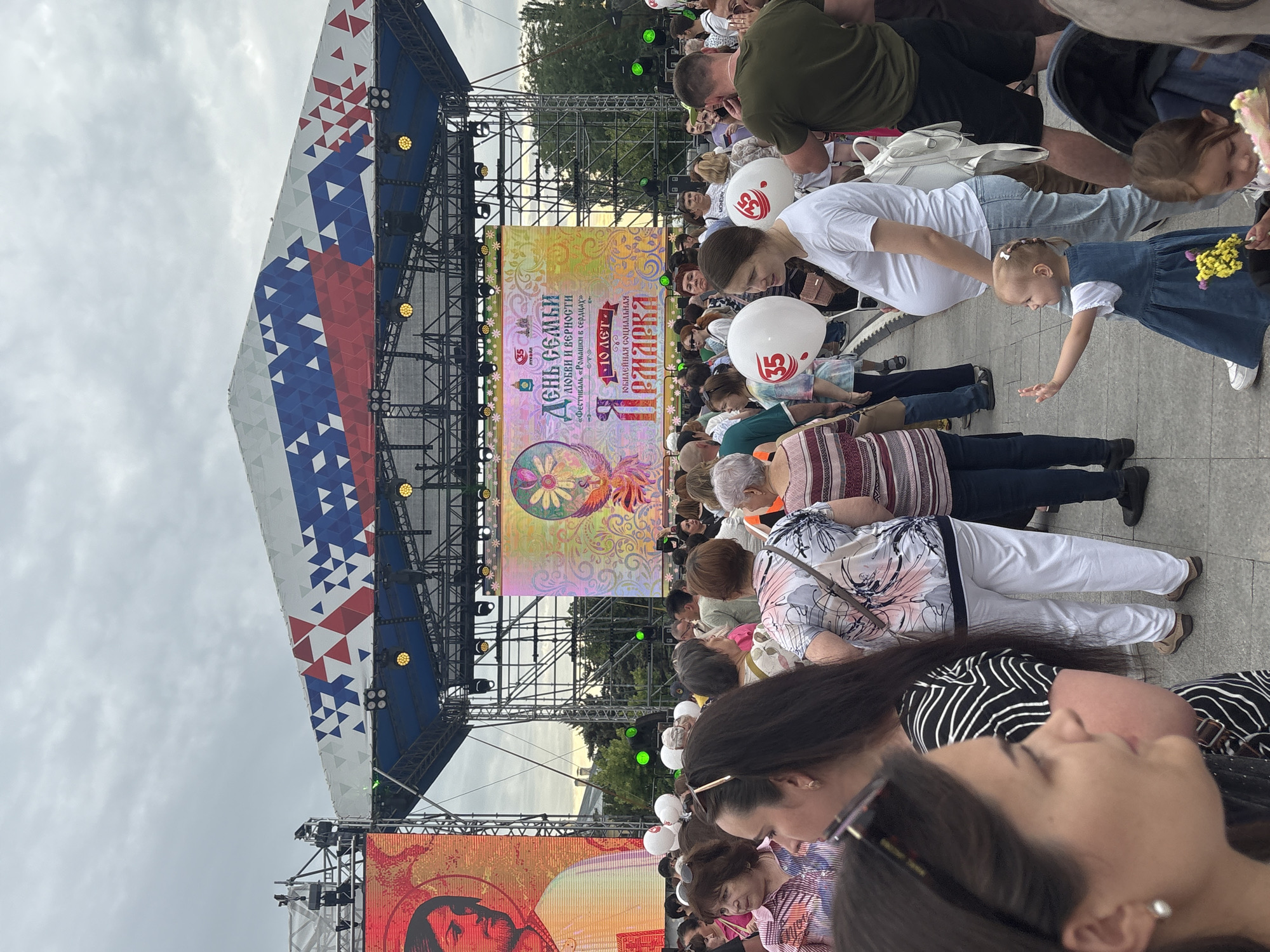 An outdoor festival is underway in Astrakhan, Russia, under an overcast sky. A large stage, adorned with a design resembling the Russian flag, dominates the background. On the stage, a large LED screen displays festive graphics and text. In front of the stage, a crowd of people, diverse in age and gender, are gathered. Many are looking towards the stage. Among the crowd, there are several individuals holding white balloons, some with the number "35" printed on them. A pregnant woman is visible, and a young child in a blue dress is gesturing with their hand. The ground appears to be paved. The overall scene suggests a public celebration or event.