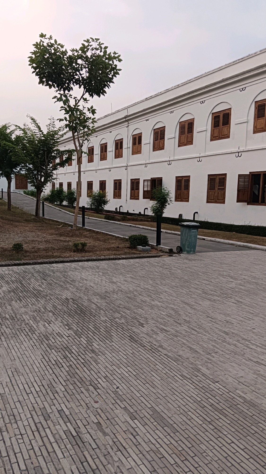 The image depicts the exterior of a white, colonial-style building with numerous arched windows featuring wooden shutters. A paved walkway leads from the foreground, which is covered in grey brickwork, towards the building. Several trees are planted along the walkway, with one tree on the left having a distinctive heart-shaped canopy. A green trash can with wheels is situated near the building's exterior wall. The sky is overcast. This building is located in Ngawi, Indonesia.
