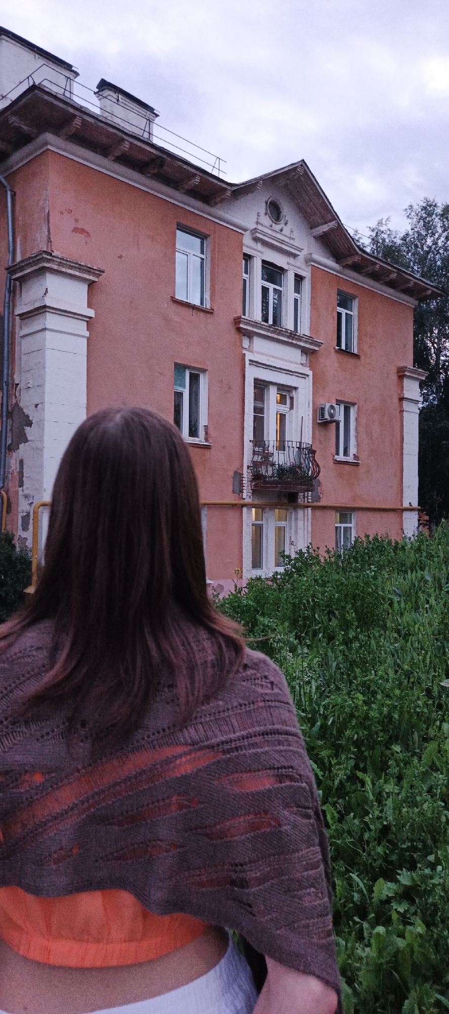 In Votkinsk, Russia, a person with long, dark brown hair stands with their back to the camera, facing a large, pink, multi-story building. The building has several windows, some with balconies. The person is wearing a brown, patterned shawl over an orange garment. Lush green vegetation surrounds the building and the person. The sky is overcast, suggesting it might be dusk or a cloudy day. The architecture of the building appears to be older, possibly from the Soviet era, with peeling paint and some visible wear and tear.