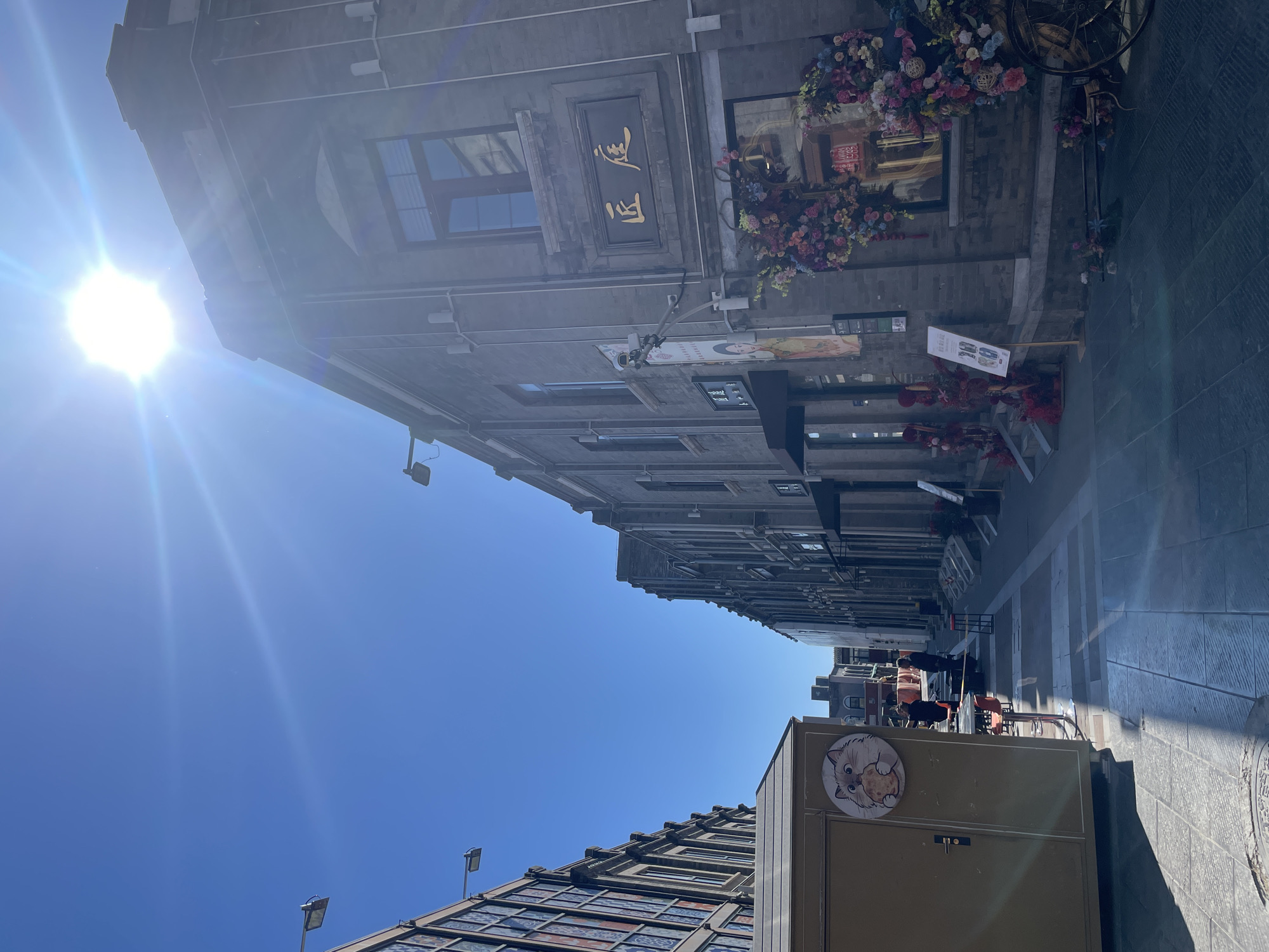 The image depicts a street scene in Dongcheng District, China, angled upwards. The perspective emphasizes the height of the buildings and the depth of the narrow street. The buildings are old, constructed from grey brick with traditional Chinese architectural elements like ornate signs and window frames. One building features Chinese characters in gold on a dark background. Floral arrangements decorate the storefronts. The street is paved with light grey stone, and there are a few people visible towards the end of the street. In the foreground, there is a brown structure with a circular logo of a cat holding a cookie. A stained glass roof structure sits behind it. A bright sun is visible in the blue sky.