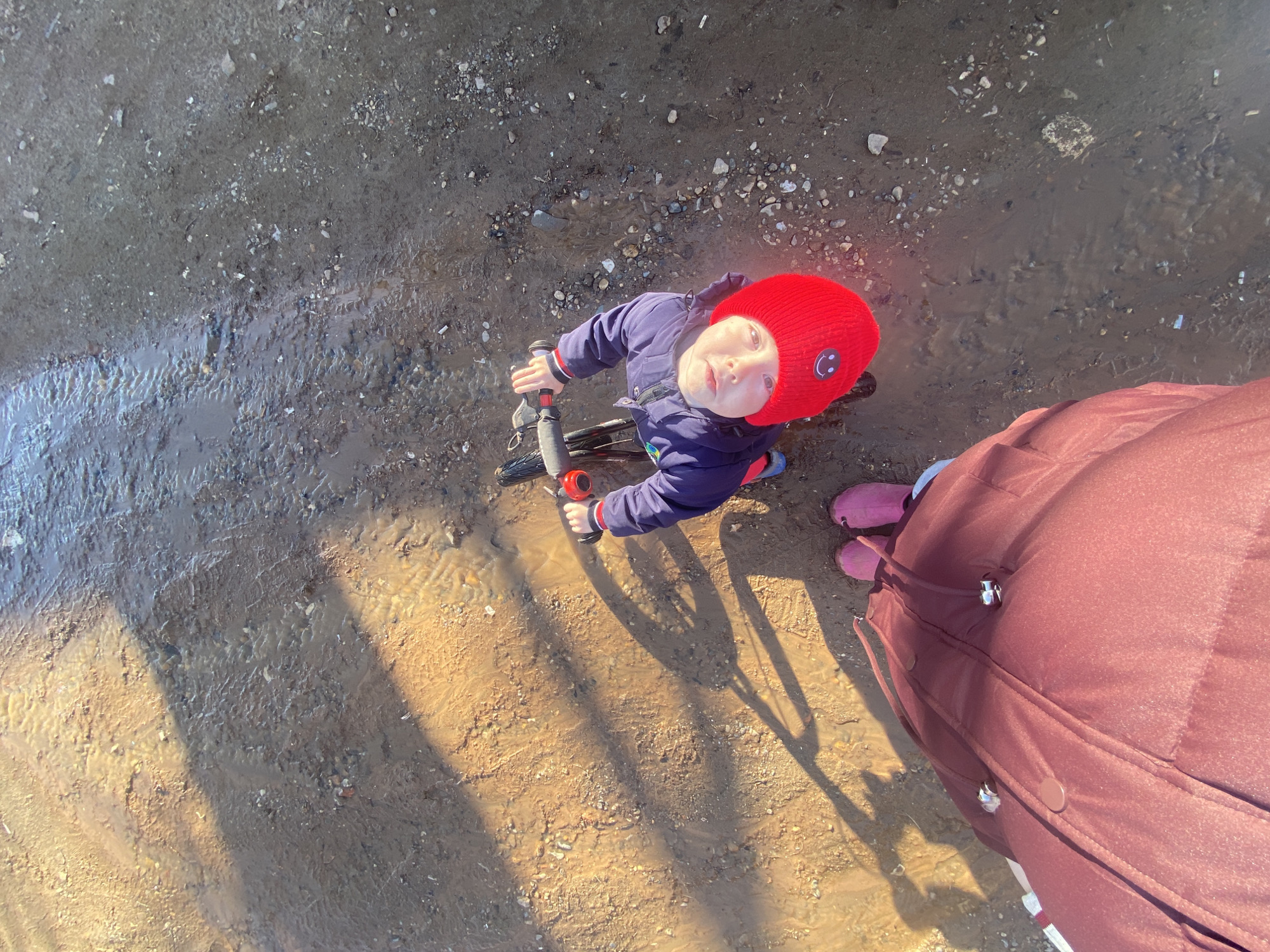 A top-down view shows a child on a bicycle standing on a muddy, watery surface. The child is wearing a red knit hat with a smiley face on the side, a blue jacket, and pink boots. A portion of a dark red coat or blanket is visible to the right of the frame. The location appears to be a dirt path or unpaved area, partially covered with water. Shadows are cast across the ground. It's likely to be Ольховка, Russia.