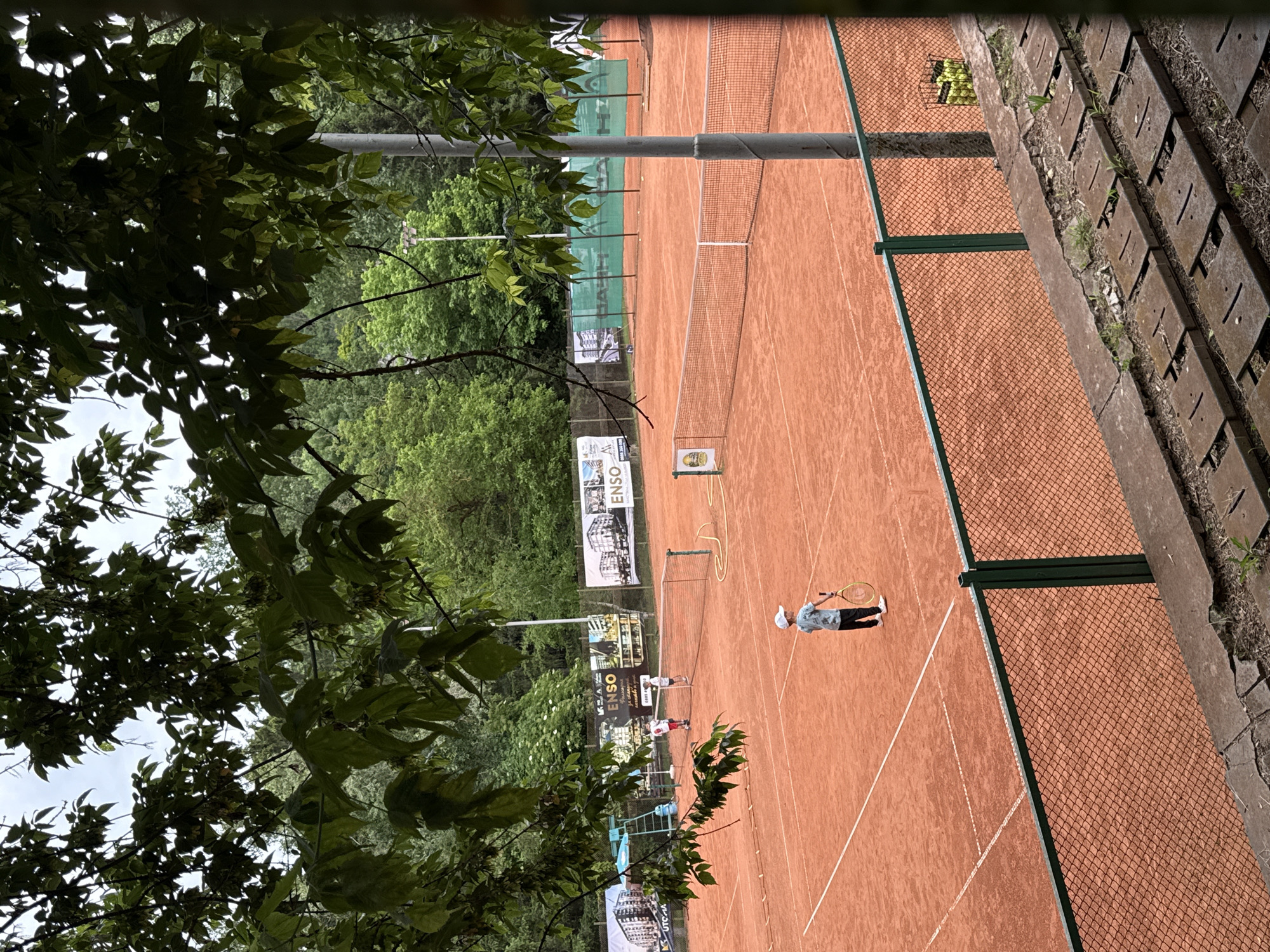 The image depicts an outdoor tennis court with a clay surface in the city of Pleven, Bulgaria. The court is framed by trees in the foreground and background. Two children are visible on the court. One child, wearing a white cap and a blue shirt, stands near the baseline holding a tennis racket. Another two children are near the net on the opposite side of the court. A tennis net divides the court. In the background, banners are displayed along the fence. A metal fence separates the spectator seating from the court. A basket of tennis balls is visible near the fence. The weather appears overcast.