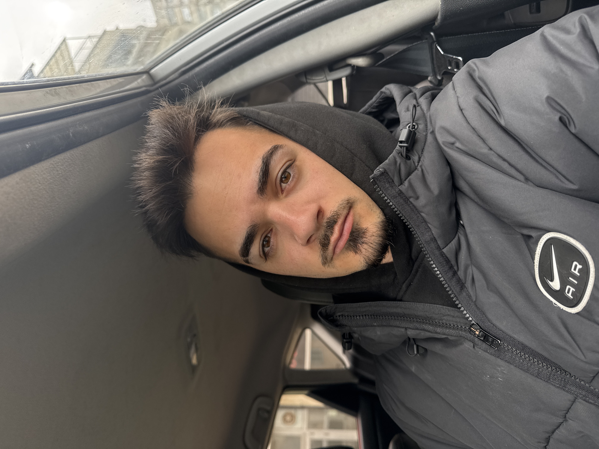 A man is posing for a selfie inside a vehicle. He has dark hair, brown eyes, and a short beard. He is wearing a black hooded sweatshirt and a black Nike AIR jacket. The interior of the vehicle is visible, including the roof and window. Outside the car, there is a building. It is impossible to determine the location in the city of Sofia, Bulgaria from the given image.
