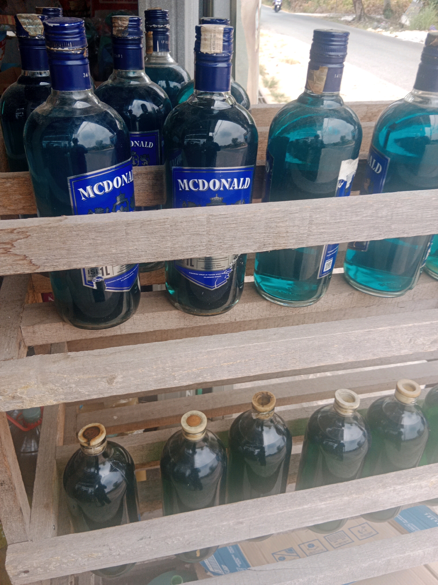 The image depicts a collection of bottles arranged on wooden shelves, likely in a shop or market stall. In the foreground, on the bottom shelf, are several dark-colored liquid-filled bottles with capped openings. The middle shelf holds larger, translucent blue liquid-filled bottles, some with blue labels that read "MCDONALD". The top shelf also contains similar blue liquid-filled bottles, with visible caps and some labels. The background shows a road and vegetation, suggesting an outdoor or semi-outdoor setting. This scene is located in the city of Banjarbaru, Indonesia.