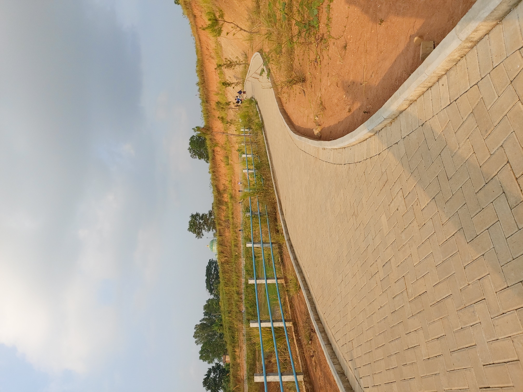 The image depicts a paved walkway curving through a park-like setting. The walkway, made of light brown interlocking pavers, is bordered by a concrete curb. To the left of the walkway, there is a body of water, likely a pond or lake, with a blue metal fence running along its edge. Beyond the water, there are trees and grassy hills. A green dome structure, possibly part of a mosque or other building, is visible on the hill in the distance. Several people are walking along the walkway and in the distance. The ground on the right side of the walkway is dry and reddish-brown earth with sparse vegetation. The sky is overcast with a mix of light and dark clouds. The scene appears to be in a public outdoor recreational area. Based on the provided context that the image is from Bandar Lampung, Indonesia, this location is likely a public park or green space within the city.
