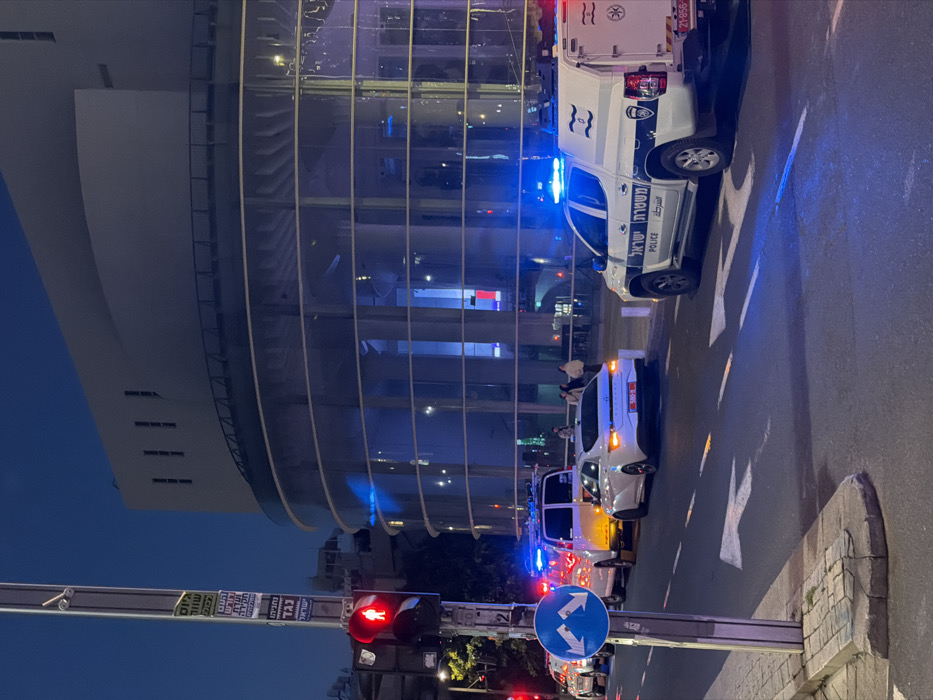 This image depicts an urban street scene at twilight or night in Tel-Aviv, Israel, specifically on Sderot Sha'ul Hamelech, in front of the Charles Bronfman Auditorium, part of the Tel-Aviv Performing Arts Center.
In the immediate foreground on the left, a metal street pole features a red pedestrian traffic light showing a "don't walk" signal, a blue circular road sign indicating "turn left or right," and several vertical placards with Hebrew text. The asphalt roadway displays white painted arrows for "straight" and "turn" lanes.
Multiple vehicles are present on the street. From left to right, appearing in a stopped or slow-moving sequence: a partially visible white emergency vehicle with active blue flashing lights, a white panel van (possibly an ambulance or support vehicle) with active amber flashing lights, a white Hyundai sedan with an Israeli license plate (580-R), and a white Israel Police pick-up truck with a utility canopy and active blue flashing lights. The police vehicle is marked with "משטרת ישראל" and "POLICE" in English, and bears an Israeli license plate (21-856-XX).
In the background, the prominent Charles Bronfman Auditorium features a large, curved glass facade on its lower sections, reflecting ambient light. The upper structure of the building is a lighter, solid material with vertical slit windows. Approximately four individuals are visible on the sidewalk in front of the building. The active emergency lights on the vehicles indicate an ongoing incident or a security presence.