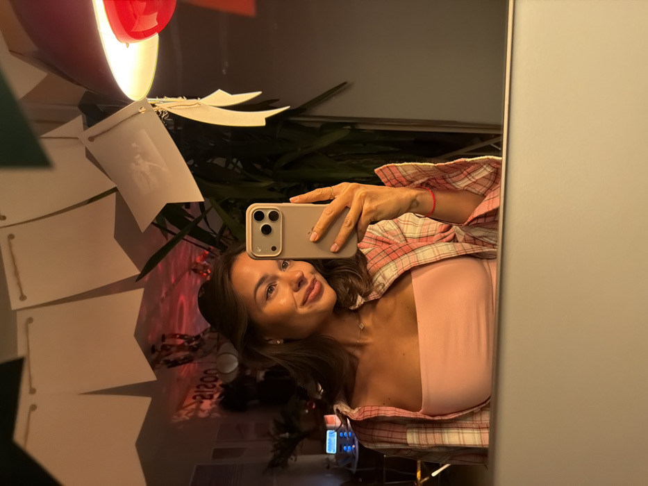 A young woman is reflected in a mirror, holding a smartphone to take a selfie. She is wearing a pink top with a plaid shirt layered on top. Behind her, there is a decorative lamp and some greenery, as well as hanging white paper decorations. The background is slightly blurred, suggesting a shallow depth of field. A reflection of a bar area can be seen in the mirror as well.